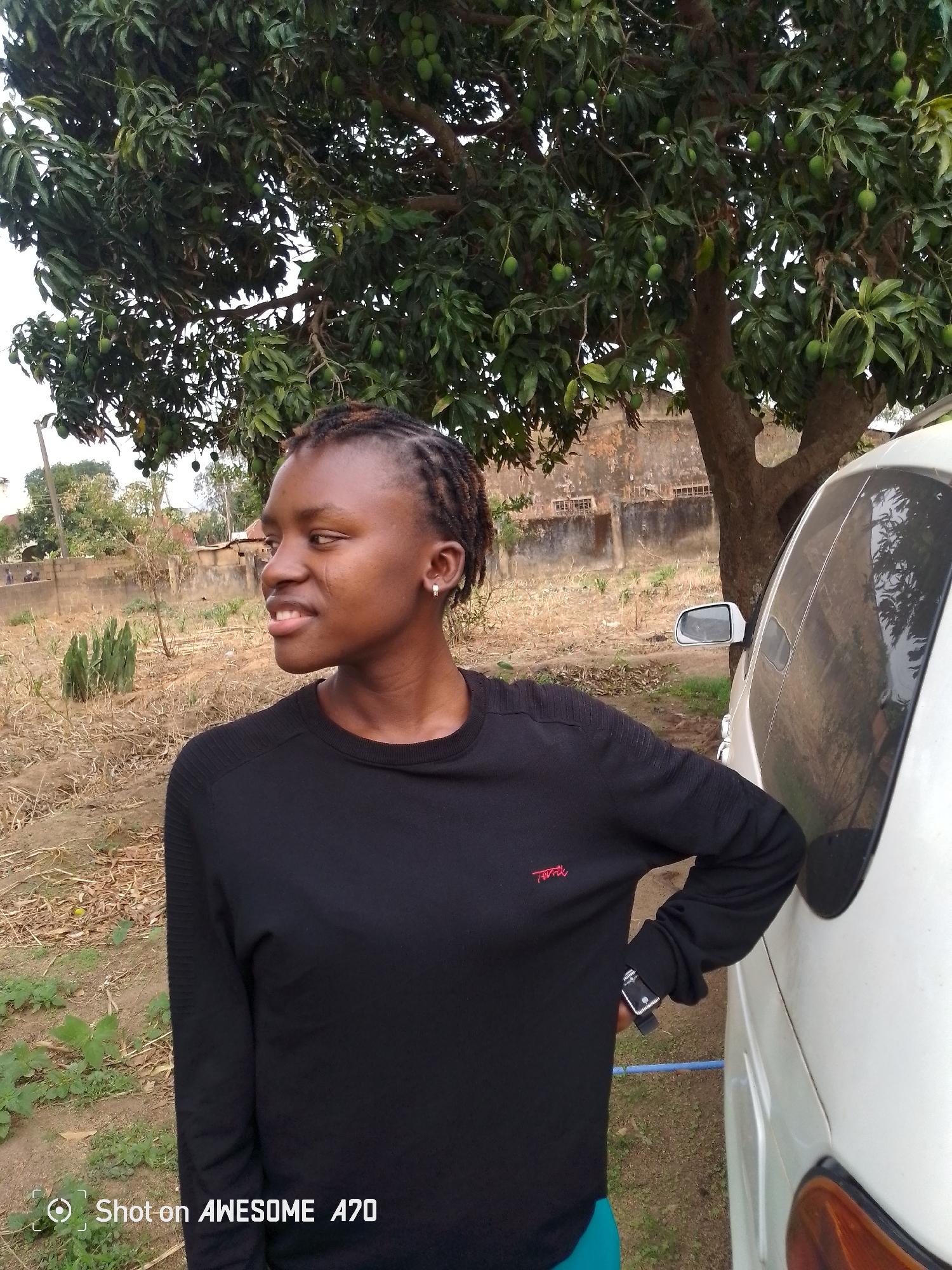 A person stands next to a white car under a tree with green fruit, likely mangoes, in an outdoor setting. The person has short braided hair and wears a black long-sleeved shirt. They are looking to the left. The car is partially visible, with its side mirror and taillight noticeable. The ground consists of dry dirt and scattered vegetation. In the background, there is a building with a fence-like structure. The location appears to be a rural or suburban area in the city of Unknown, Unknown. The photograph was taken with an "AWESOME A70" phone.