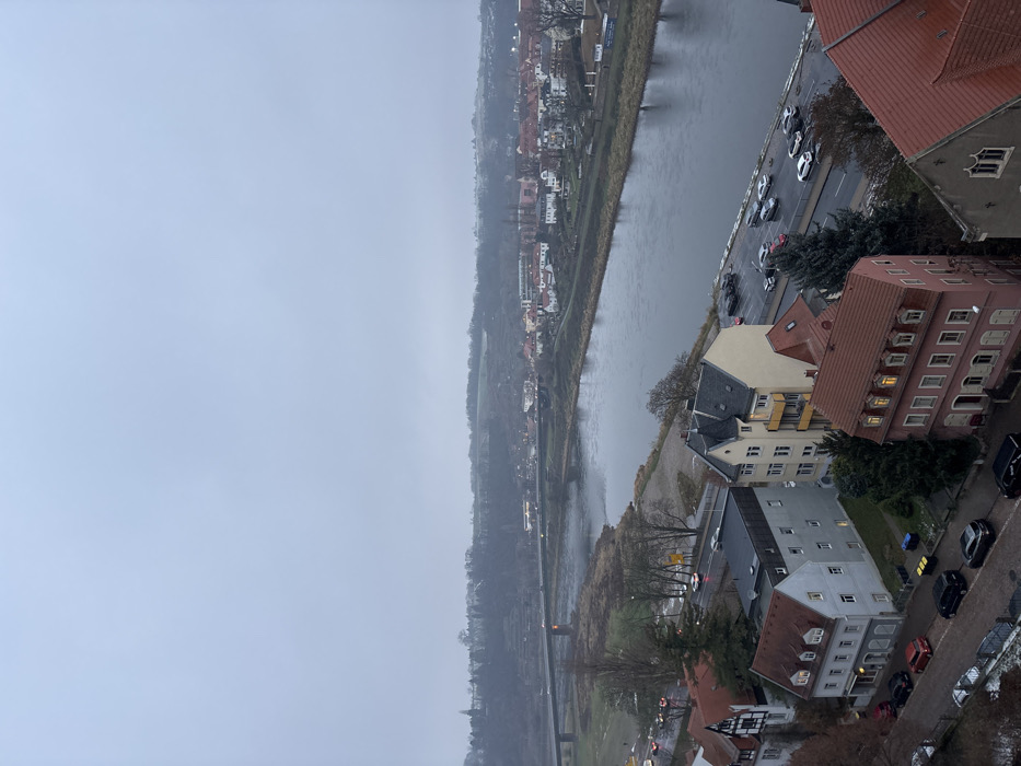 The image captures an elevated, panoramic view of a town or city, likely Meissen, Germany, overlooking a river on a heavily overcast day. The perspective is from above, looking down towards the river and the landscape beyond. The sky is a uniform, pale grey, indicating a completely cloudy day with low light conditions, possibly late autumn or winter due to the bare trees.
A wide river, appearing calm and reflecting the grey sky, flows from the foreground towards the distant horizon, creating a prominent horizontal element across the middle of the scene. Along the near bank of the river, a paved road runs parallel to the water. Numerous cars are parked along this road, and some appear to be driving, though no individual people are clearly visible. A collection of buildings, primarily multi-story structures with varying architectural styles, some featuring red-tiled roofs and others darker, flat roofs, line this near bank. Warm lights are visible in some windows, suggesting either late afternoon or interior lighting due to the dim day.
On the far side of the river, another line of buildings, smaller and more interspersed with vegetation, follows the riverbank. Beyond these structures, a tree-covered hillside rises, showing patches of dull green and brown, extending into the misty distance where the landscape fades into the grey sky. A bridge with visible arches spans the river further downstream (towards the left of the image). The overall atmosphere is muted and somewhat somber, characterized by the grey tones of the sky, river, and damp-looking ground. No discernible text is visible.