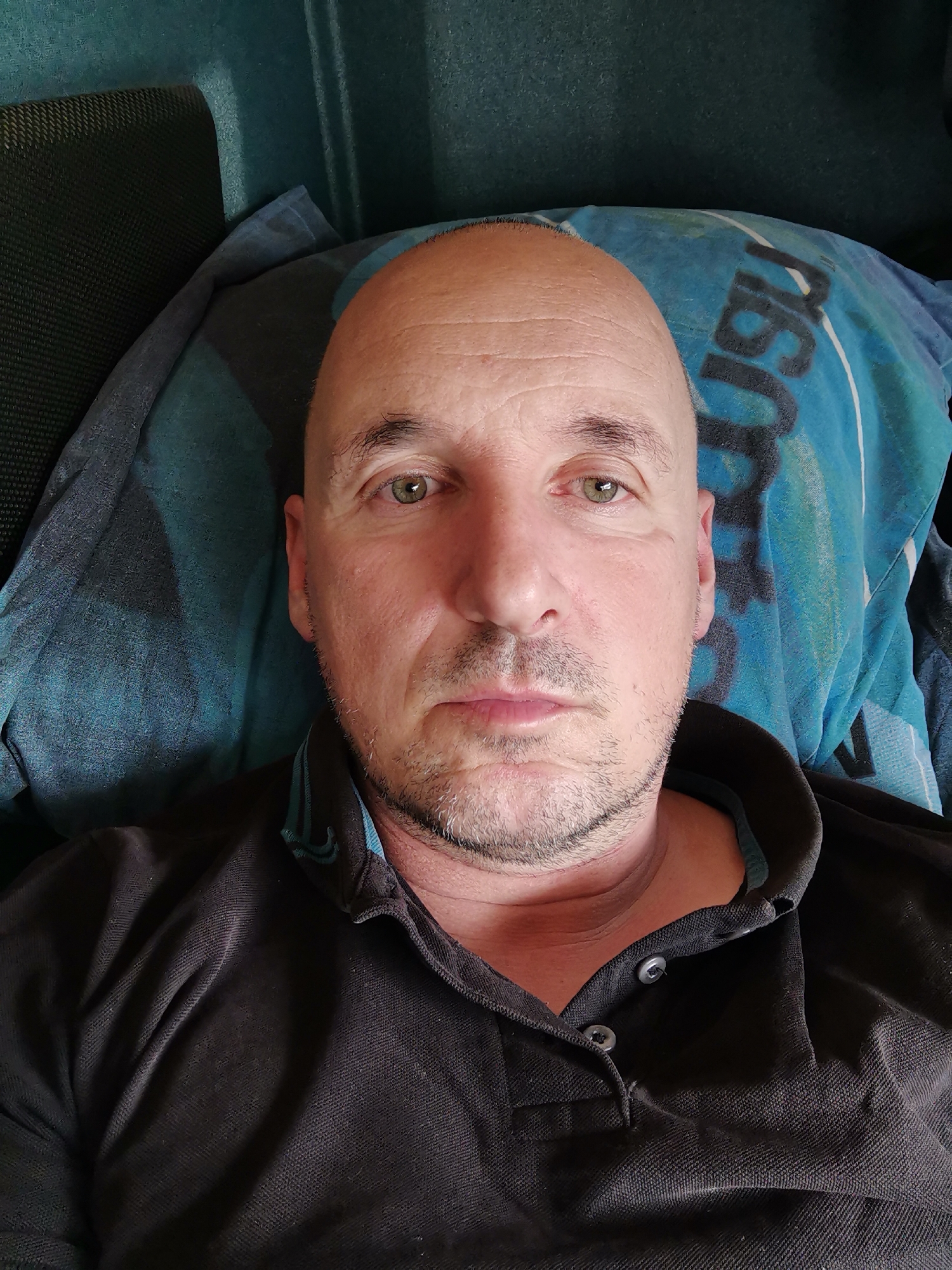A bald, middle-aged man with light green eyes and stubble is lying down. He is wearing a black, button-down shirt and is positioned against a blue pillow or bedding with a dark print. The background suggests an interior setting, possibly a bed or couch. The location in the city of Karapelit, Bulgaria is not evident from the provided visual information.