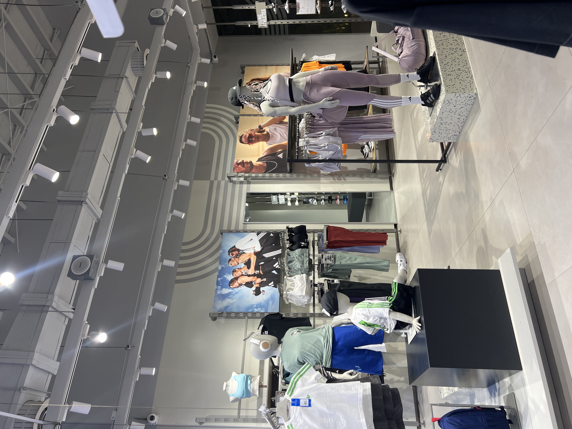 This media file captures an indoor retail setting, likely a sportswear store given the merchandise and branding. The scene is dominated by product displays and mannequins, showcasing athletic apparel.
Several mannequins are positioned throughout the store. In the foreground, a female mannequin is dressed in a lavender sports bra and matching leggings with white stripes, wearing black sneakers and a cap with braids. Her display stand is a speckled grey and white block. Another section features mannequins wearing t-shirts, shorts, and sports bras in various colors such as light green, blue, and white, with some displaying Adidas branding. One mannequin, wearing black shorts and a white t-shirt with green stripes, is positioned on a black display cube.
The walls feature large promotional posters showcasing diverse models wearing Adidas sportswear. One poster shows a female and male model; another shows several male models, all actively posing in athletic gear.
The store's interior design is modern and bright, with a primarily grey and white color palette. The ceiling has exposed structural elements and numerous white track lights illuminating the displays. The flooring consists of light-colored tiles. A small white security camera is visible on the ceiling.
Visible text includes a sign that reads "YOUNG ATHLETES" above one of the clothing racks. Adidas logos are also discernible on clothing tags and the large wall posters. No actual people are present in the immediate view, and there is no active event taking place beyond the static retail display.