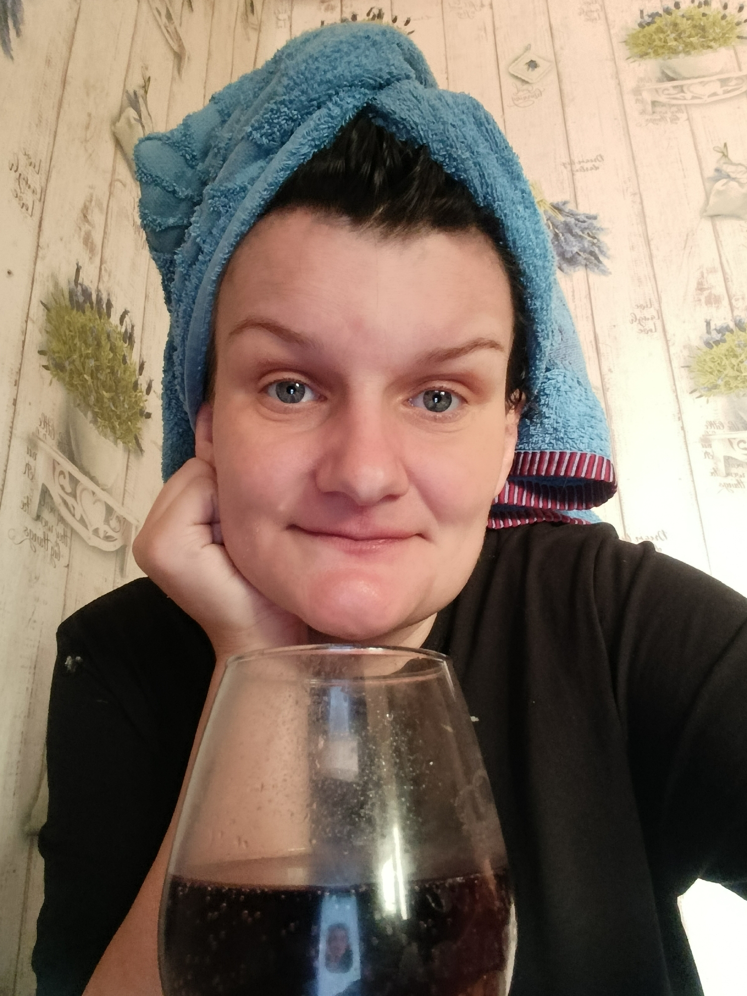 A woman with blue eyes and dark hair wrapped in a blue towel rests her head on her hand. A wine glass filled with a dark liquid is in the foreground, obscuring part of her body. She is wearing a black shirt. The background is a wall with a floral pattern. The location of Фоки, Russia, cannot be confirmed from the image.