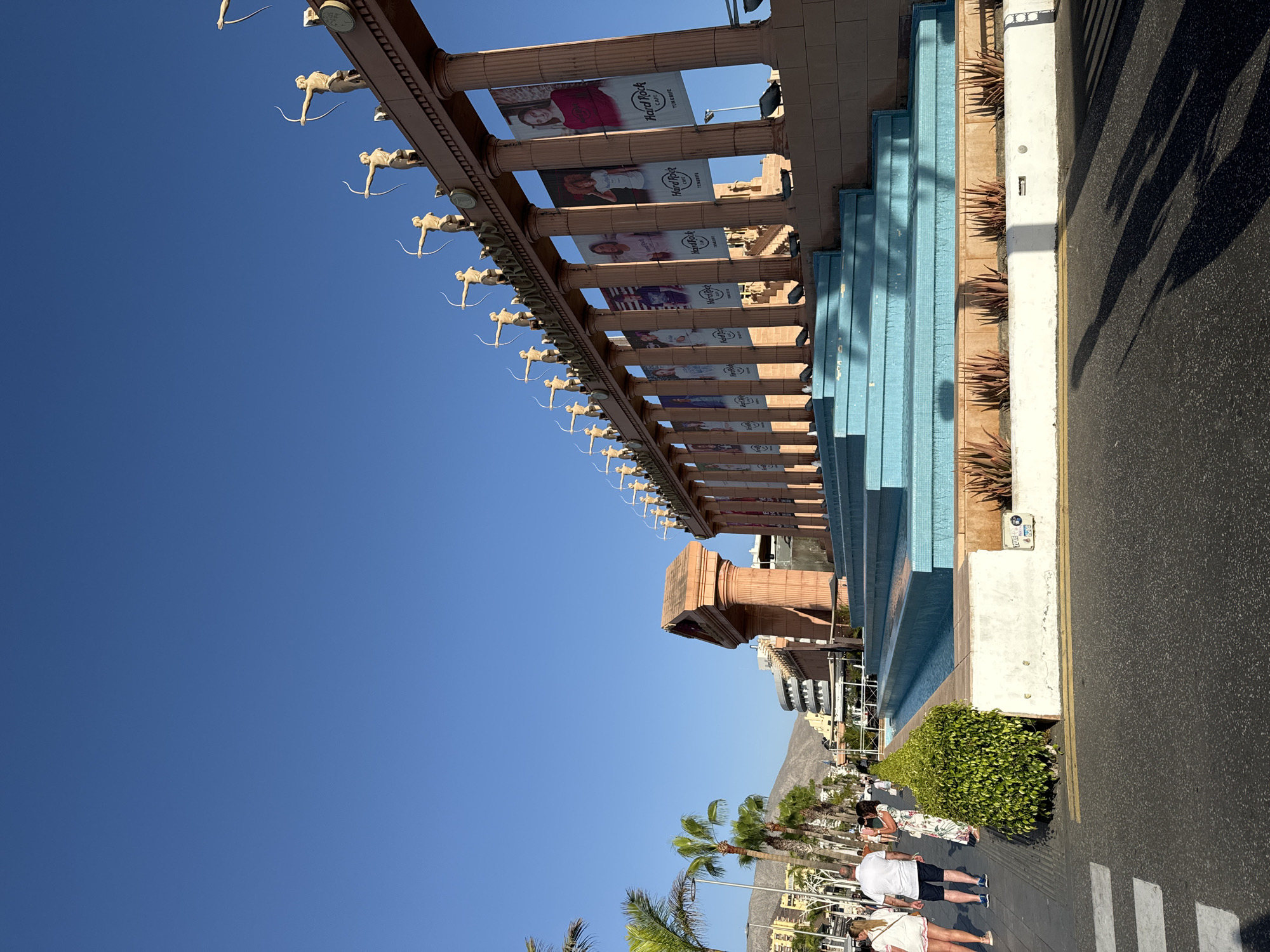This image captures the Plaza del Duque in Arona, Spain, on a clear, sunny day. The foreground shows a street with a pedestrian crossing and shadows of people walking. To the right, there is a large, ornate structure with a colonnade and statues of archers along the top. Several banners, featuring the Hard Rock Cafe logo, hang between the columns. In front of the colonnade, there is a tiered blue fountain with several levels. Beyond the fountain, there are lush green bushes and further into the background, there are buildings, palm trees, and mountains under a bright blue sky. On the sidewalk to the left, several people are walking, dressed in casual summer clothing. One woman in a white dress and another in a white top and shorts are visible. A man in a white polo shirt and shorts is also present, as is a woman in a floral dress. The overall scene depicts a lively public space, likely a tourist area, with a blend of modern and classical architectural elements.