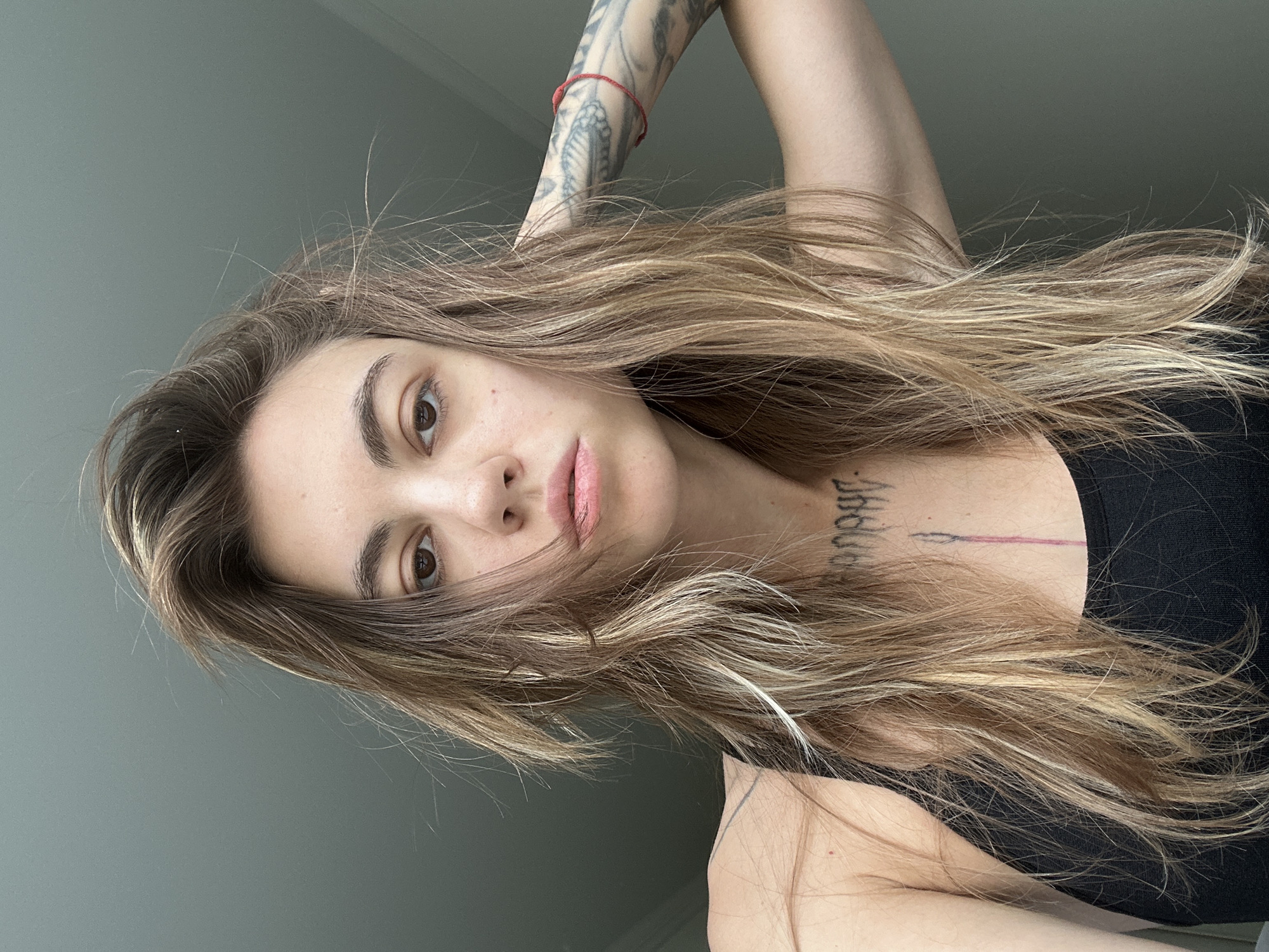 The image is a close-up, selfie-style shot of a young woman with long, wavy, light brown hair with blonde highlights. She is wearing a black tank top. The woman has tattoos on her arm and chest. She is looking directly at the camera with a neutral expression. The background is a plain, light gray wall. There are no discernible landmarks or features that indicate a specific location within Moscow, Russia.