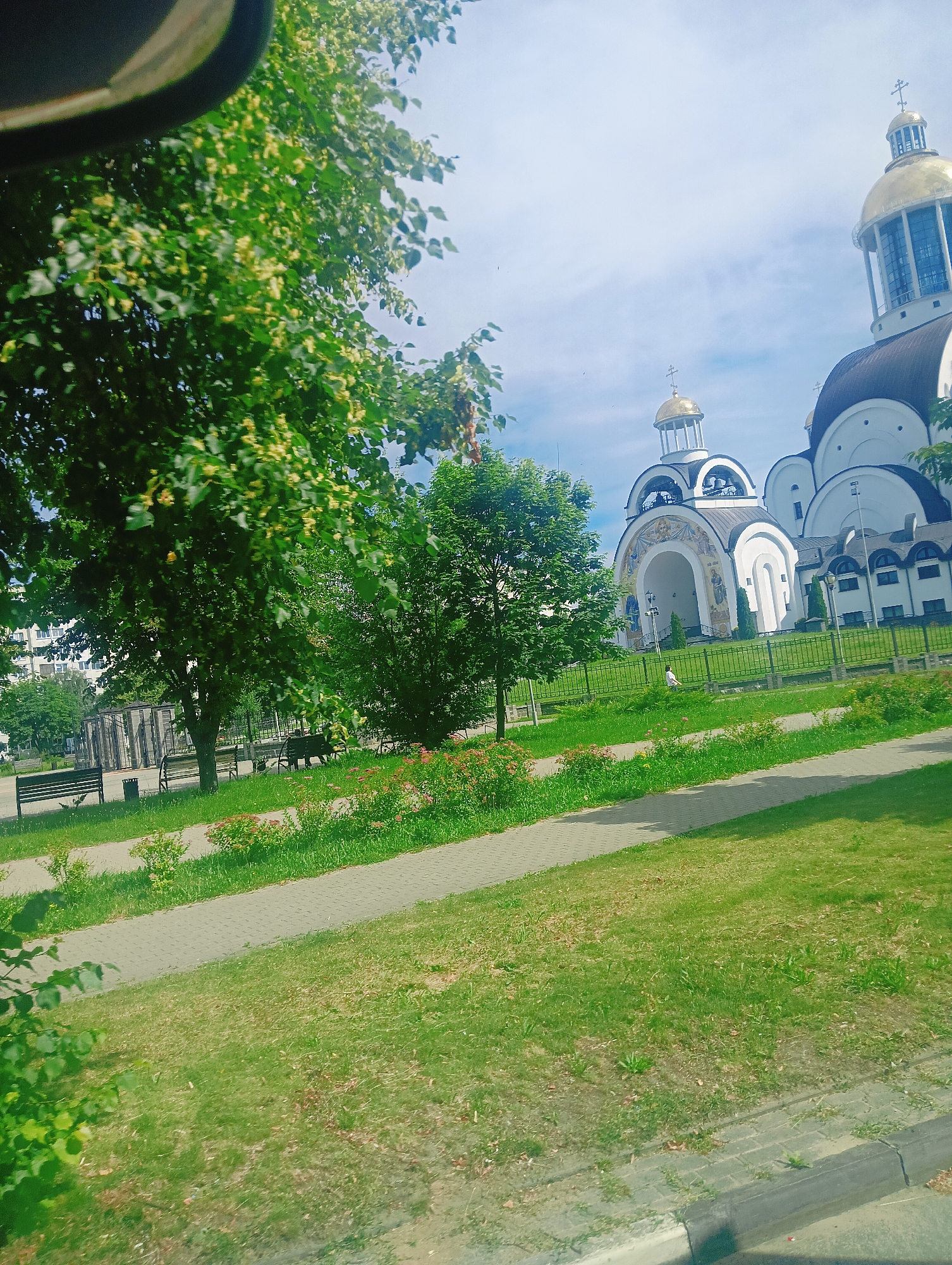 The image depicts a bright, sunny day in Salihorsk, Belarus. The scene is a park area adjacent to a large, white Orthodox church with a prominent golden dome and several smaller domes. Green trees with lush foliage, some bearing small yellow flowers, frame the view, with the leaves of a large tree in the foreground casting dappled shadows. A paved walkway winds through the park, bordered by grass and flowerbeds with pink blooms. In the distance, behind a black metal fence, a few people can be seen near the church. Several dark-colored benches are placed in the park. The overall atmosphere is peaceful and serene, suggesting a pleasant outdoor environment. The time of day is likely late morning or early afternoon, given the bright sunlight and clear sky.