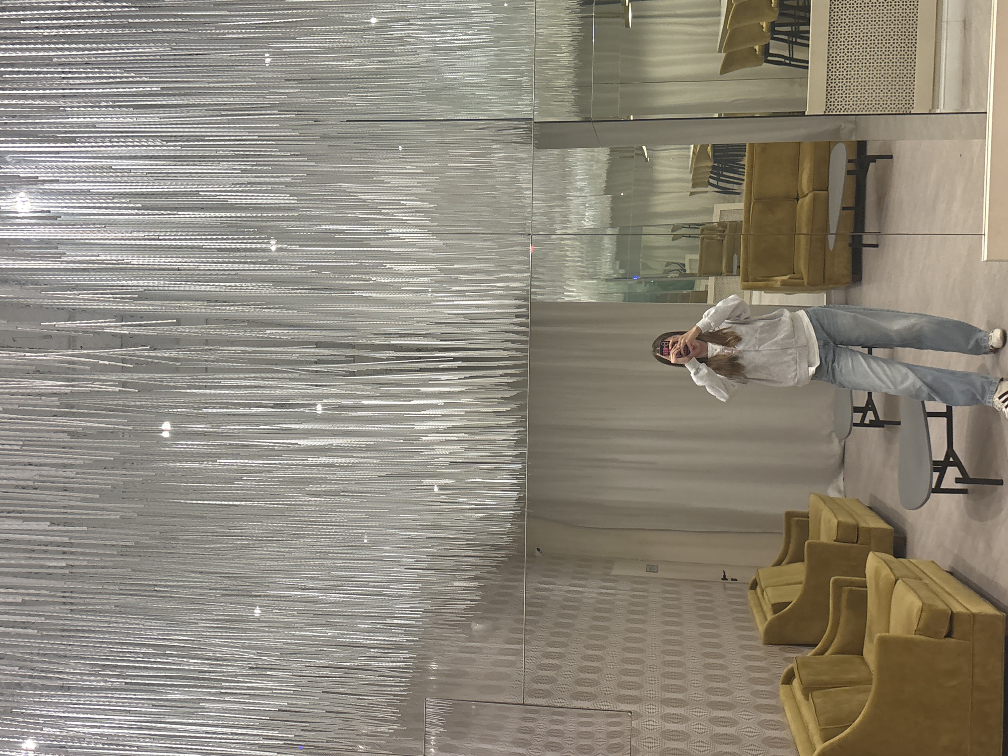 A young woman stands in what appears to be a showroom, possibly in the city of Perm, Russia. She is taking a selfie in front of a mirrored wall, capturing her reflection along with the surrounding environment.
The woman is wearing a light-colored hoodie or sweatshirt, blue jeans, and Adidas sneakers. She is holding her phone up to take the photo, and her face is partially obscured by the phone.
Behind her and reflected in the mirror, there are several mustard-colored chairs or small sofas arranged in rows. Small round tables stand next to the furniture. The floor is covered in a patterned carpet.
To the left of the mirror, there is a large wall installation composed of numerous thin, horizontal lines, possibly decorative rods or lighting elements. The installation occupies a significant portion of the image and adds a distinctive visual element to the scene.