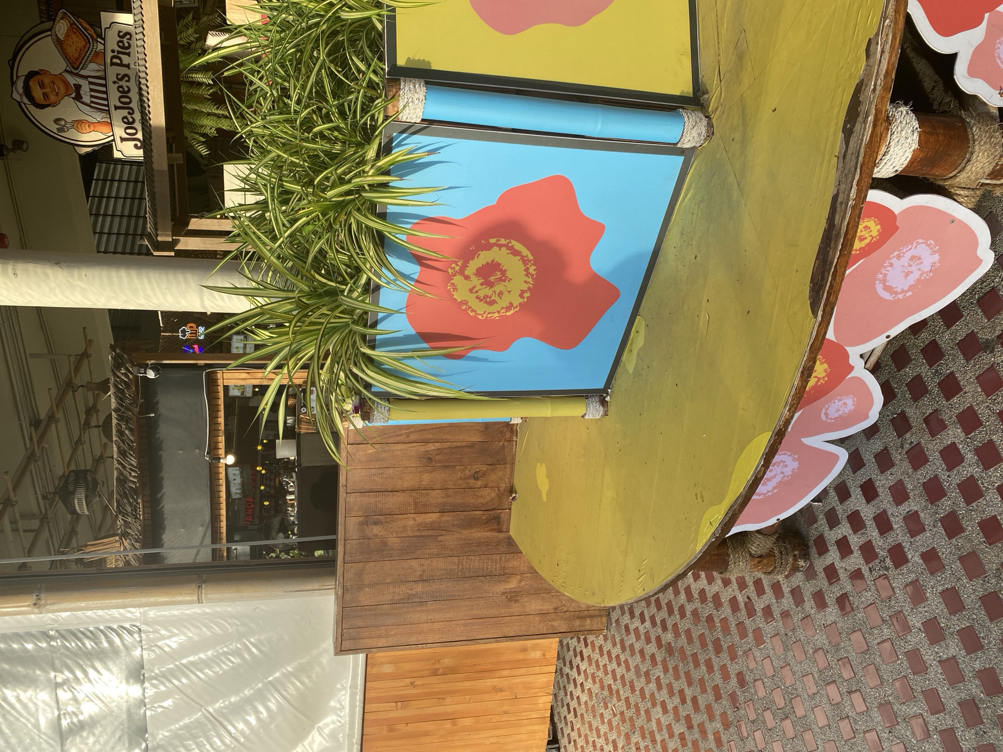 The image shows an outdoor seating area in front of JoeJoe's Pies in Baan Chaweng, Thailand. A round, yellow table is in the foreground, decorated with floral motifs. Behind the table, a planter filled with green foliage has panels with colorful flower designs. In the background, a sign for "JoeJoe's Pies" is visible. The ground is paved with reddish-brown square tiles. The overall scene suggests a casual dining or relaxation spot.