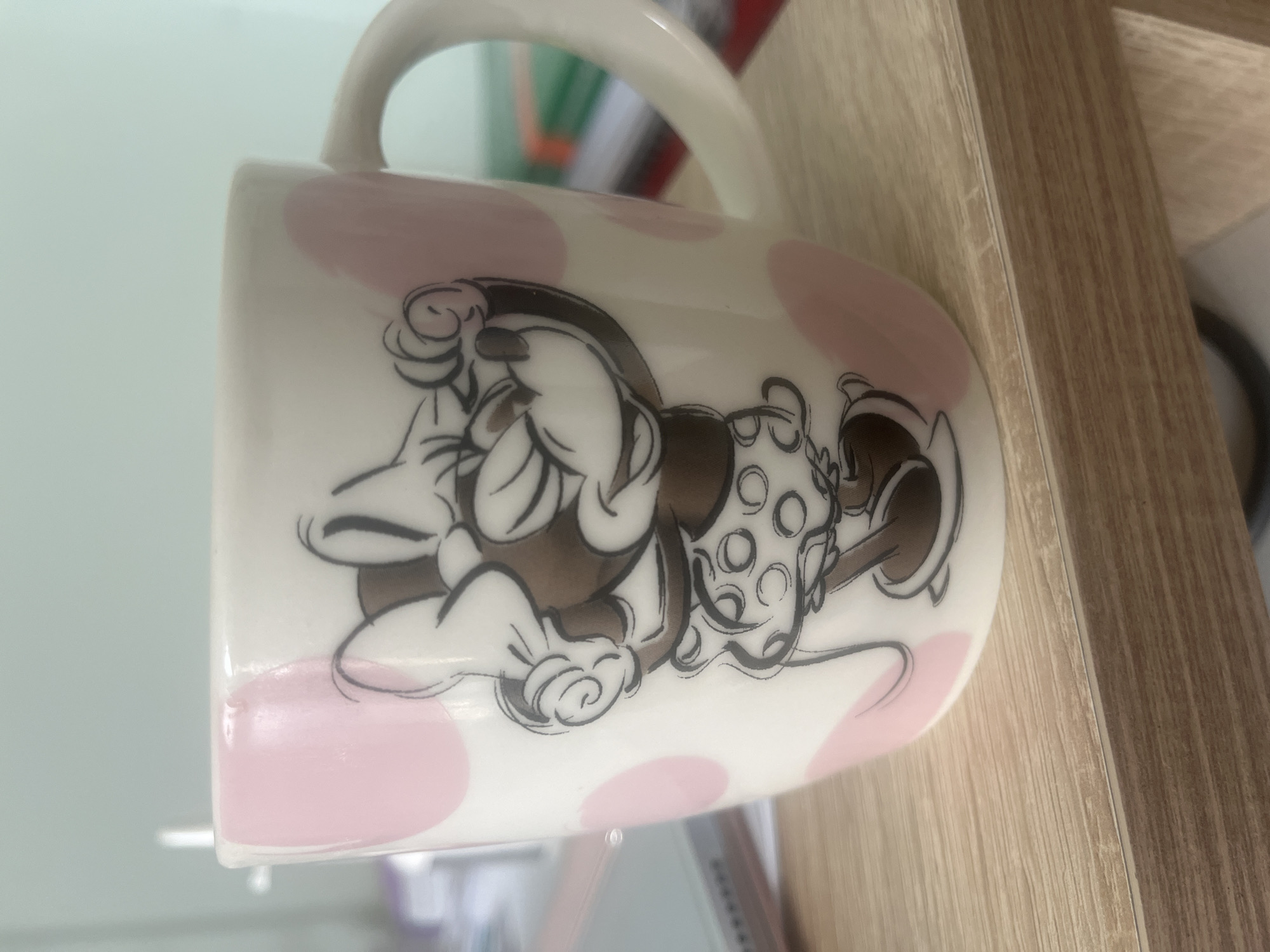 The image shows a white mug with pink polka dots and a cartoon illustration of Minnie Mouse, placed on a light brown wooden surface. The mug is slightly tilted. It is unclear whether the location of the image is in Plovdiv, Bulgaria.