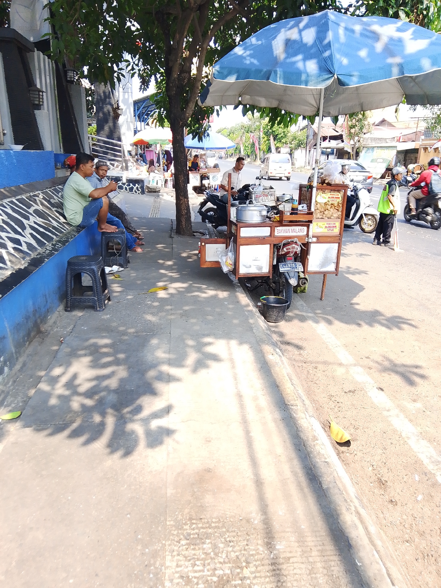 The image depicts a street scene in Potroyudan, Indonesia, on a sunny day. In the foreground, a paved sidewalk runs alongside a street. Several black plastic stools are placed on the sidewalk, with two people sitting on a raised blue and white tiled bench to the left. A street food cart, adorned with a blue umbrella, is positioned on the sidewalk. The cart displays food items and menus, with a license plate visible on its rear. A person in a yellow vest stands near the cart, holding a broom. In the background, vehicles, including motorcycles and cars, are visible on the street. Trees and buildings line the street, and shadows from foliage are cast on the sidewalk, indicating the presence of overhead trees. The overall environment suggests a bustling urban setting with ongoing commercial activity.