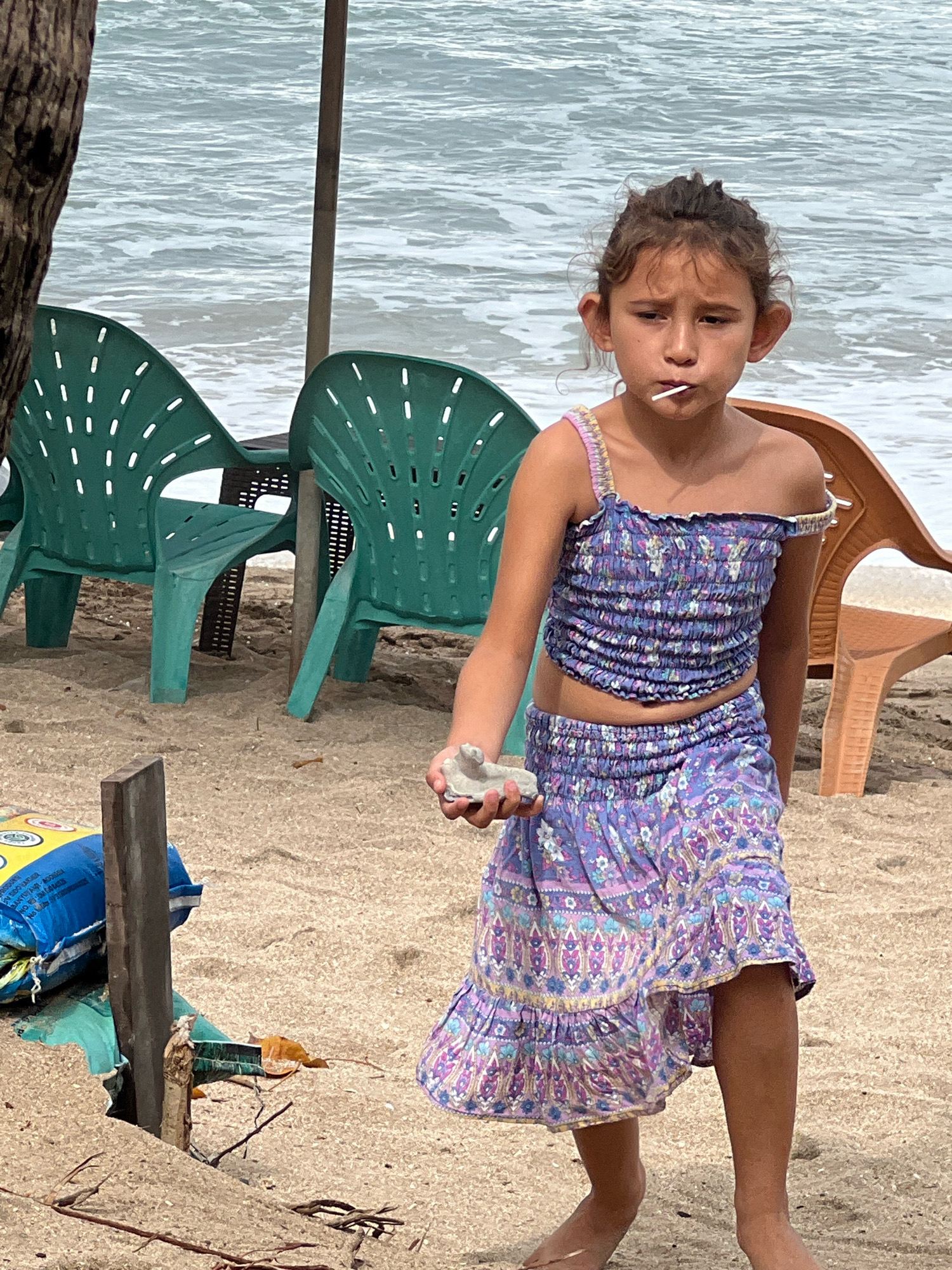 A young girl with curly brown hair and large ears stands on a sandy beach in Kuta, Indonesia, holding a light gray object in her right hand and a lollipop in her mouth. She is wearing a purple floral print two-piece outfit, consisting of a smocked crop top and a ruffled skirt. The background shows the ocean with moderate waves, and several empty green plastic chairs and one brown plastic chair are positioned on the sand. A brown wooden pole is visible in the foreground, and a blue bag with yellow lettering is partially obscured behind it.