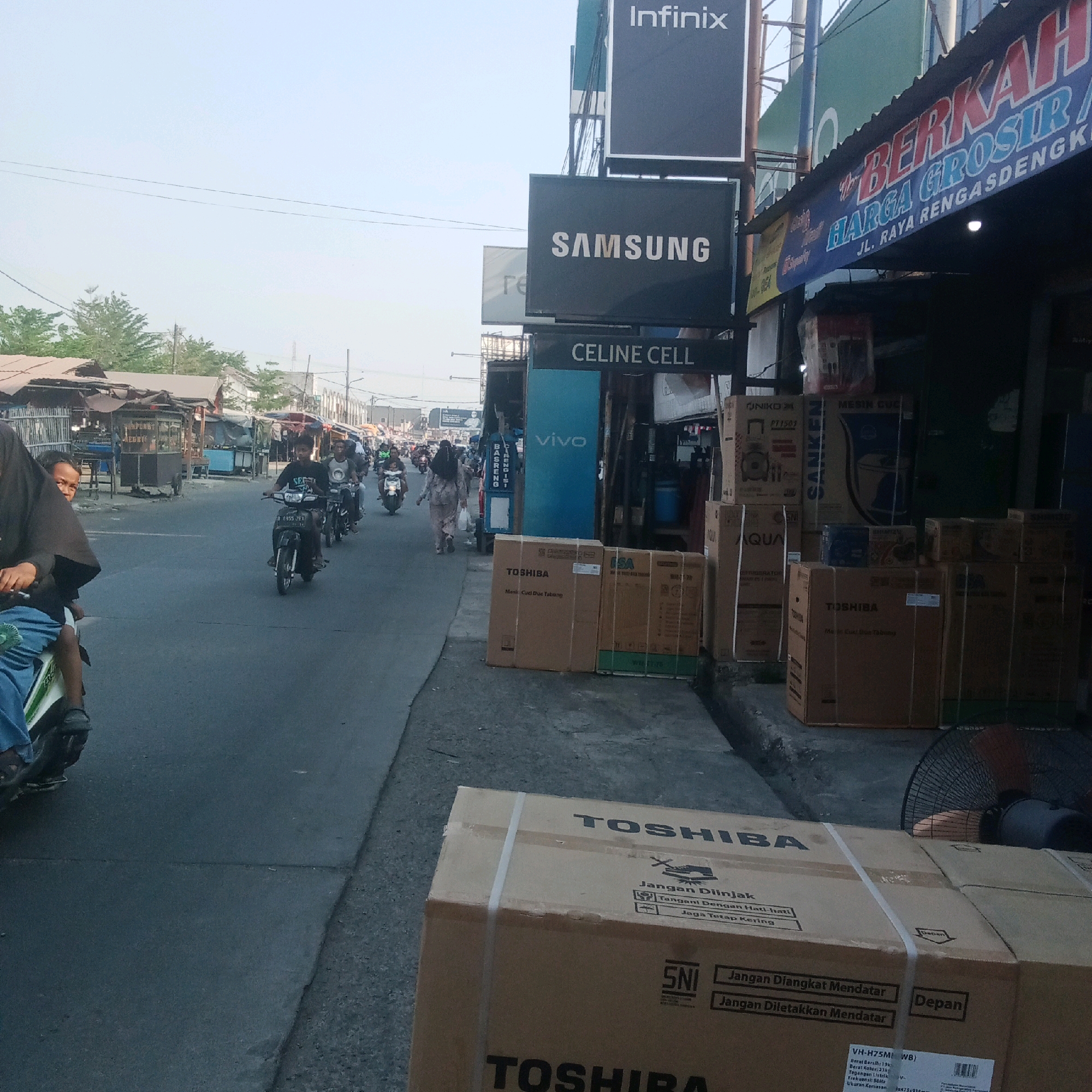 The image depicts a street scene in Karawang, Indonesia, likely a commercial area. A paved road with a slight incline runs through the center, with a sidewalk and market stalls on the right. Several people are on motorbikes, moving along the road. On the right side of the road, there are shops and stalls displaying various products, including electronics like Samsung, Infinix, and Celine Cell. Several large cardboard boxes, some labeled "Toshiba" and "Aqua," are stacked on the sidewalk, suggesting retail or delivery activity. A fan is visible next to the stacked boxes. The sky is bright, indicating daylight, and the environment appears to be a busy, possibly humid, outdoor market.