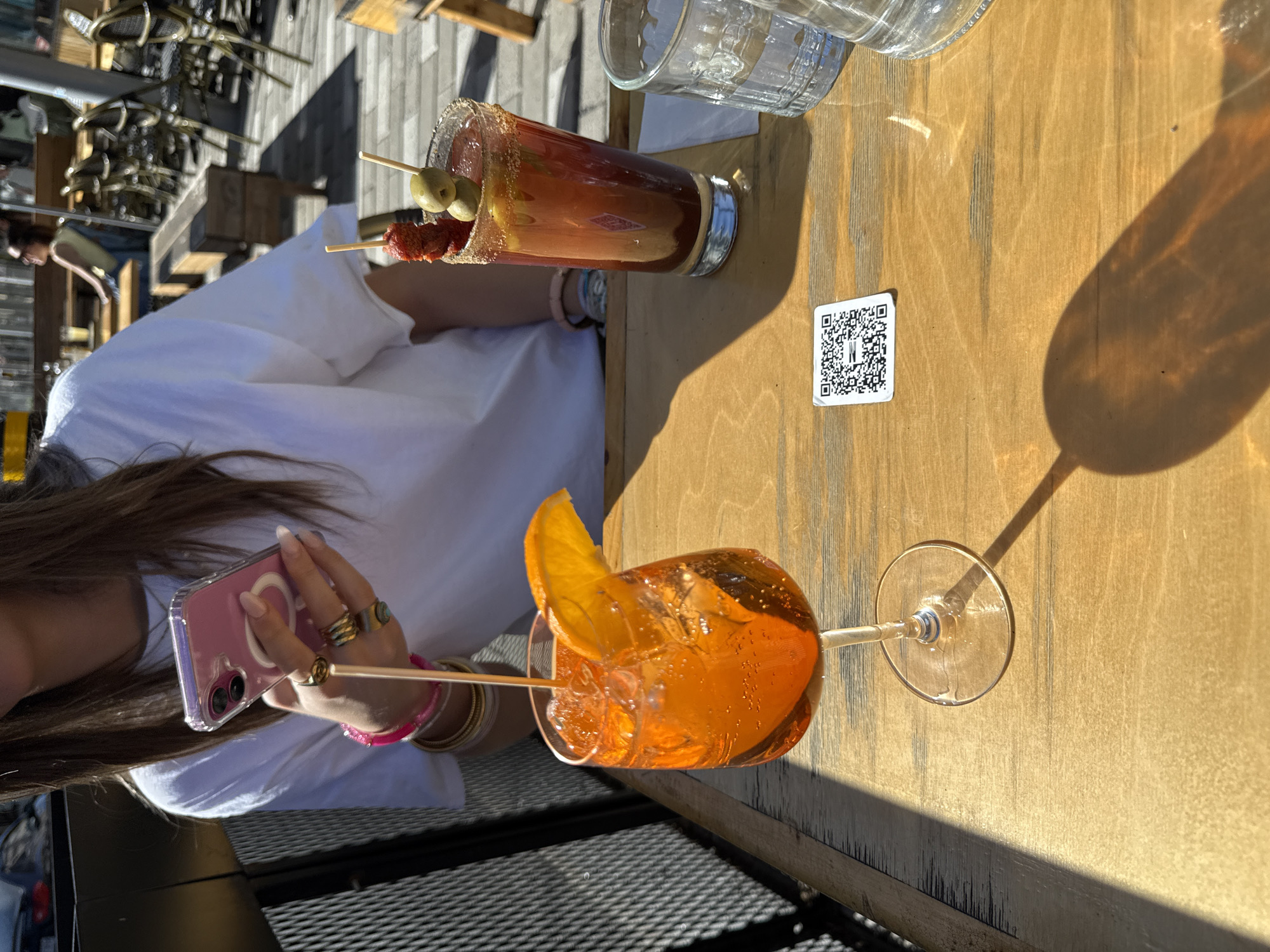 This outdoor scene, set in Montreal, Canada, captures a relaxed moment on a sunny day. The primary subjects are two colorful cocktails and a person partially visible in the foreground.
On a light wooden tabletop, two drinks are prominently featured. To the left, a tall glass holds a deep red beverage, likely a Bloody Mary, distinguished by a spiced rim and garnished with two green olives and pieces of what appears to be cured meat on a skewer. Beside it, a clear glass of water stands. Further right, a stemmed wine glass contains a vibrant orange drink, consistent with an Aperol Spritz, garnished with a fresh orange slice and filled with ice. Strong shadows cast by the glasses indicate bright sunlight, suggesting a mid-day or afternoon setting.
A person with long brown hair, wearing a white top, is visible in the lower-left corner, holding a pink-cased smartphone with a visible MagSafe ring and camera lenses. Their hand is adorned with multiple rings, including one with a turquoise-like stone, and several bracelets.
On the wooden table between the drinks, a small white QR code sticker is visible, featuring a black pattern and a white "W" in its center. The background shows an outdoor patio setting with overturned chairs stacked, suggesting a restaurant or bar environment, and a patterned ground surface. The overall atmosphere is leisurely and bright.
