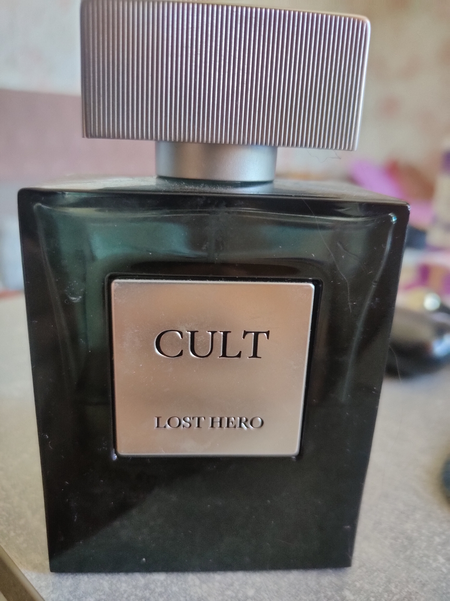 The image shows a close-up, eye-level, indoor shot of a black glass perfume bottle with a silver cap. The bottle has a silver rectangular label with raised text that reads "CULT" at the top and "LOST HERO" at the bottom. The background is blurred and appears to be an indoor setting with a light-colored wall and some out-of-focus objects. The image quality is fair, with some dust or smudges visible on the bottle and cap. The location of this image in Salavat, Russia, is not discernible from the visual content.