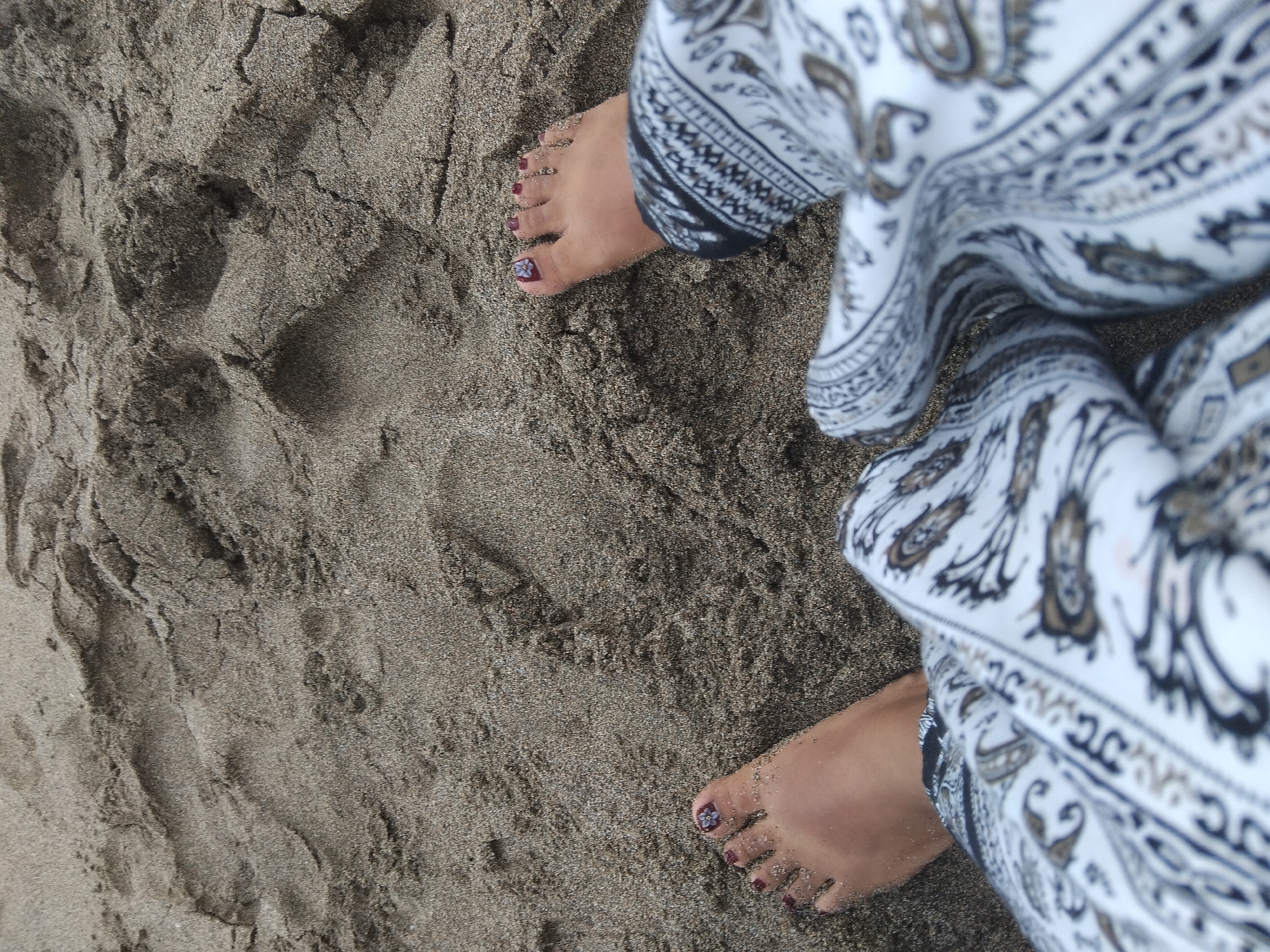 A high-angle close-up shot shows a person's feet partially buried in sand on a beach. The sand is dark brown and uneven, with footprints visible. The person's toes are visible, with dark red nail polish and a small purple flower embellishment on the big toe of the left foot. The person is wearing loose-fitting white pants with a black and brown paisley pattern. The scene is likely located on a beach in North Kuta, Indonesia.