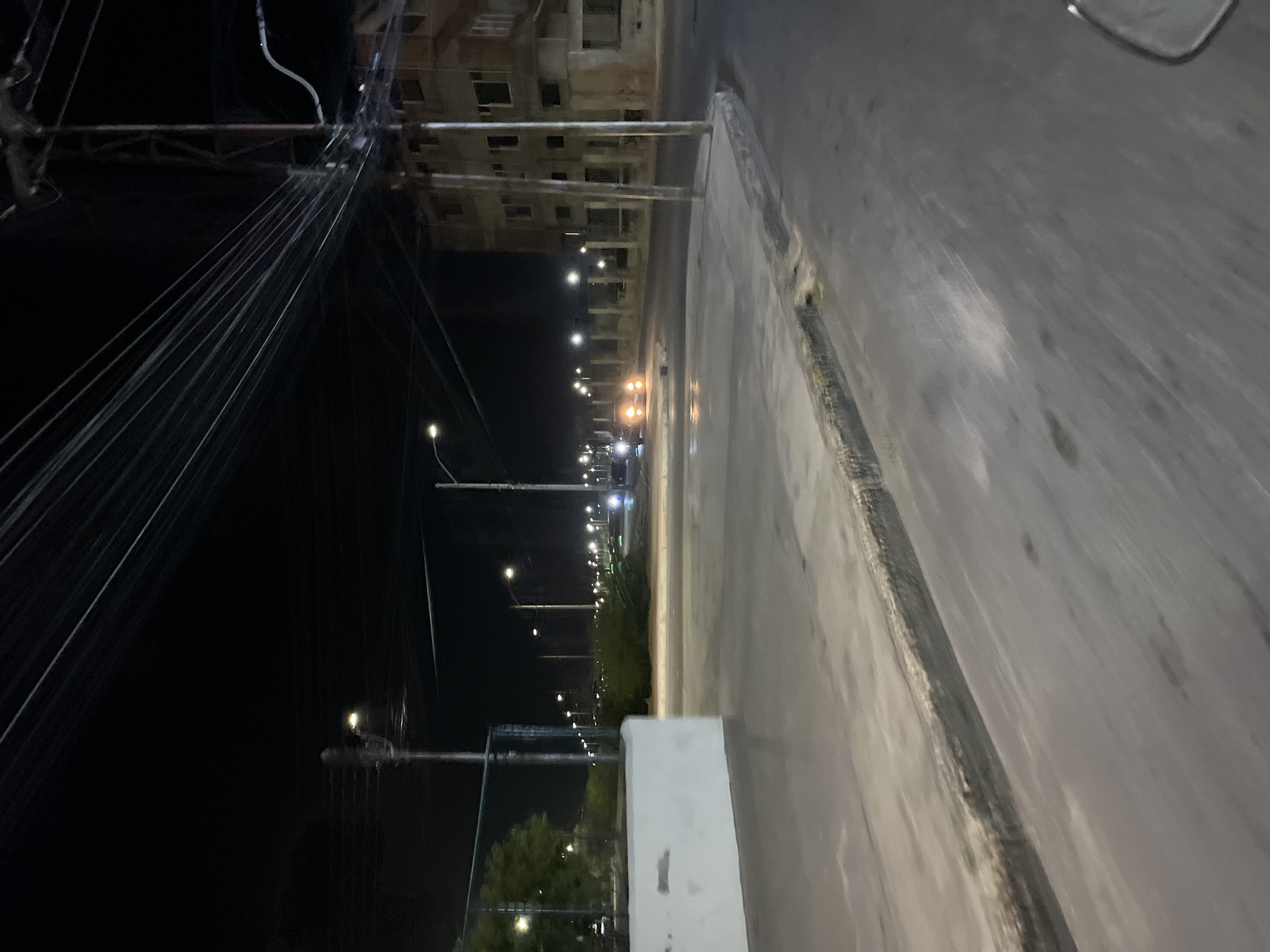 The image depicts a nighttime scene on a road in Juan Griego, Venezuela. A multi-story building, possibly under construction, is visible on the left side of the road. Streetlights line the road, extending into the distance. Power lines are tangled in the foreground, obscuring parts of the building. The right side of the image shows the paved road surface.