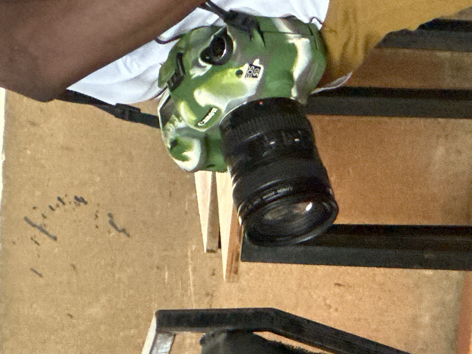 An image, oriented vertically, depicts a person holding a digital SLR camera in a close-up, slightly downward-angled perspective. The person's dark-skinned arm and part of a white shirt are visible in the upper left of the frame. The camera body is covered in a distinctive green, black, and white camouflage pattern, designed to resemble military camouflage. A black lens is attached to the camera, pointing towards the lower right.
Notable text on the camera includes "Canon" embossed on the front grip area and a label on the side reading "EOS 5D," indicating the camera model. The lens also has faint text, likely "Canon" and other technical specifications, though not fully legible.
In the background, a textured, light brown surface, possibly a wall or floor, is prominent. Dark, horizontal bar-like structures are visible in the mid-ground and lower portion of the image, suggesting a railing, shelf, or framework. Some light-colored wooden planks or supports are also seen near the camera lens. The lighting appears even, without harsh shadows.
The scene captures a moment of a person handling photographic equipment, possibly preparing for a shot or reviewing images. The setting appears to be a functional space, possibly indoors or a semi-enclosed area within Lagos, Nigeria. No specific activity beyond holding the camera is clearly depicted, nor are emotions or time of day discernible.