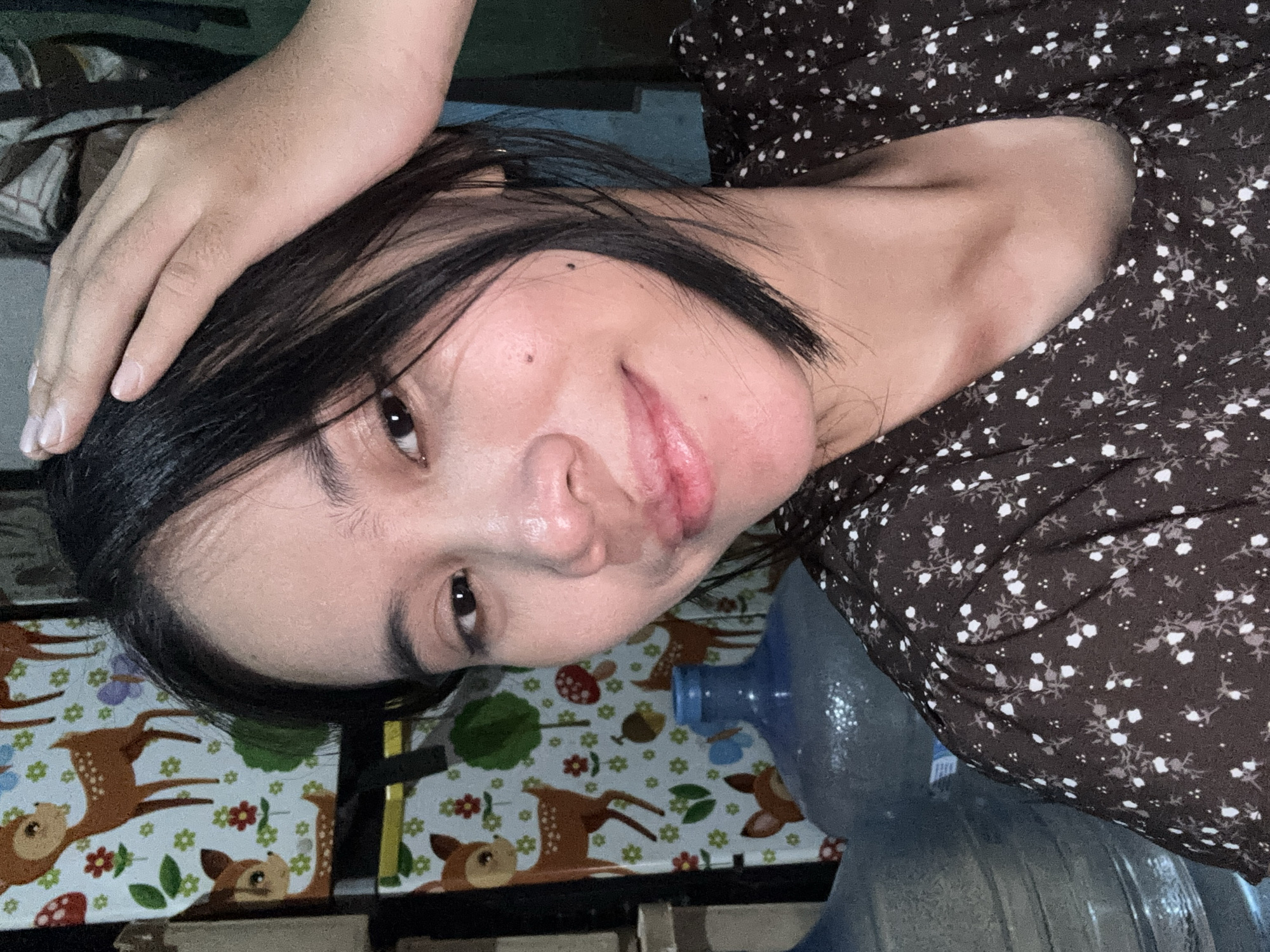 A young woman with dark hair is pictured in a selfie. She is wearing a dark brown dress with a white floral pattern. Her right hand is touching the top of her head. She is looking at the camera with a slight smile and her lips are slightly parted. The background is a colorful wallpaper with deer, flowers, and mushrooms. To the left of the woman, there are several large water cooler jugs. The overall scene appears to be an indoor residential setting in East Jakarta, Indonesia.