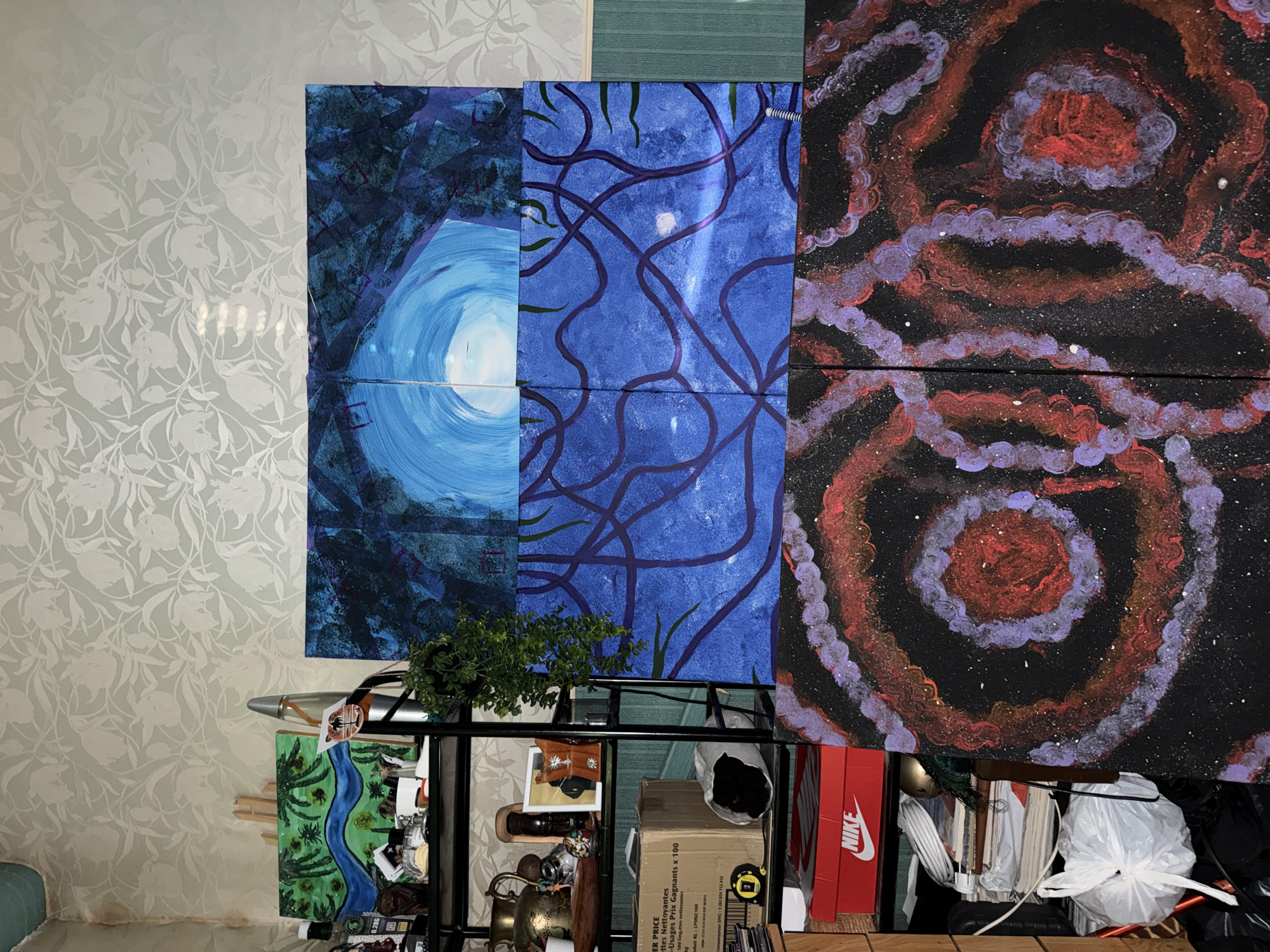 The image shows a collection of paintings and various objects arranged on and around a black metal shelving unit. The paintings, displayed on top, include a blue abstract artwork with a swirl motif, a blue painting with dark purple lines, and a dark painting with concentric rings of red and purple. Below these, on the shelves, is a smaller landscape painting featuring a river, along with various miscellaneous items such as a potted plant, a Nike shoebox, cleaning wipes, and other objects in a cluttered arrangement. The background consists of a patterned wallpaper on the left and a plain green surface on the top. The location is likely indoors in a residential setting, but there is no information available to determine the exact location in the city of Domont, France.
