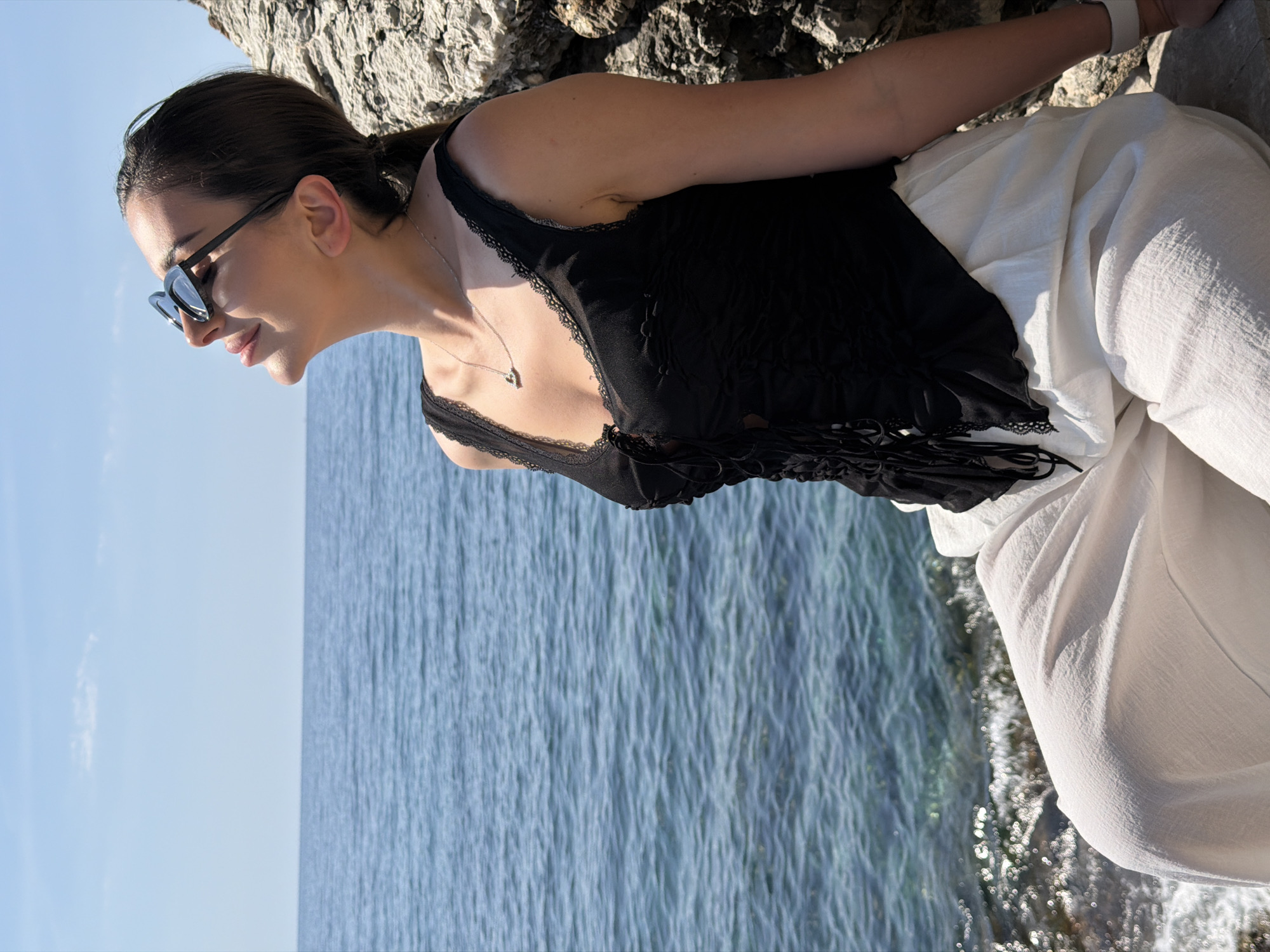 A medium-shot, portrait-oriented image shows a woman posing outdoors, leaning slightly forward and looking downwards, with a gentle smile. She is wearing dark sunglasses, a black top with lace trim and a tie-front detail, and light-colored (likely white) wide-leg trousers. A delicate silver chain necklace with a small, light-colored pendant is visible around her neck. Her hair is dark brown, pulled back, and she has fair skin.
The setting appears to be a sunny day by a coastal area, likely Budva, Montenegro, given the context. Behind her is a bright, clear blue sky on the left side of the frame, transitioning into a body of blue-green, rippling water on the right. The water shows varying shades of blue and green, indicating clarity, and some sunlight reflecting off its surface. The background also includes rough, textured rock formations, suggesting a cliffside or rocky shore, against which she appears to be leaning or standing near. The sunlight casts subtle shadows, indicating a bright, clear day. There is no visible text in the image.