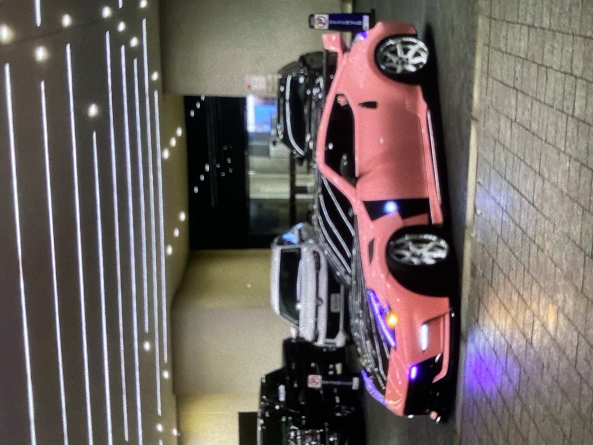 The image shows a pink sports car parked on a brick-paved surface. It is aligned with several other vehicles in a parking area, possibly near a building with illuminated vertical light fixtures. The specific location within Lhokseumawe, Indonesia cannot be determined from the provided visual information. There are no people visible in the frame.