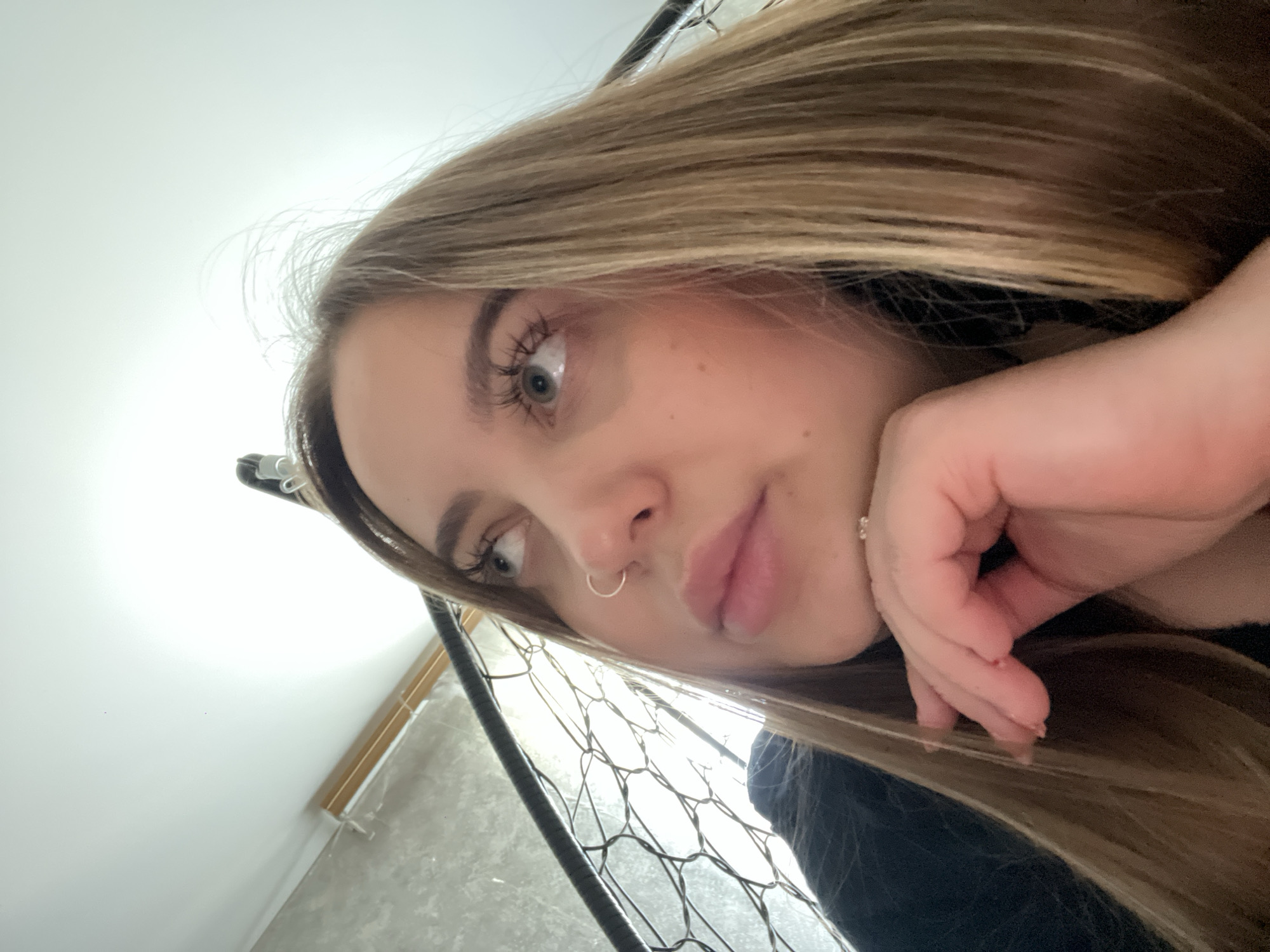 A close-up portrait shows a young woman indoors in Novopokrovskaya, Russia, captured from a low, tilted angle. She has long, light brown or blonde hair that falls around her face and shoulders. Her light-colored eyes, possibly blue or green, are visible, with prominent eyelashes, and she gazes to her left. She wears a small, silver-toned hoop nose ring and a subtle ring on a finger of her left hand, which rests near her chin or cheek in a relaxed pose. Her lips are full with a natural or light pink tone. She is wearing a dark-colored top.
The background features a brightly lit, plain white wall or ceiling dominating the upper half of the frame, suggesting a well-lit indoor environment, likely during the daytime. The lower part of the background shows a dark, open-weave mesh or net structure, possibly part of furniture like a bed frame or a decorative element. Below this, to the right, are partially visible light-colored wooden blinds or a window frame. The woman's expression appears neutral or slightly contemplative. No text is visible in the image.