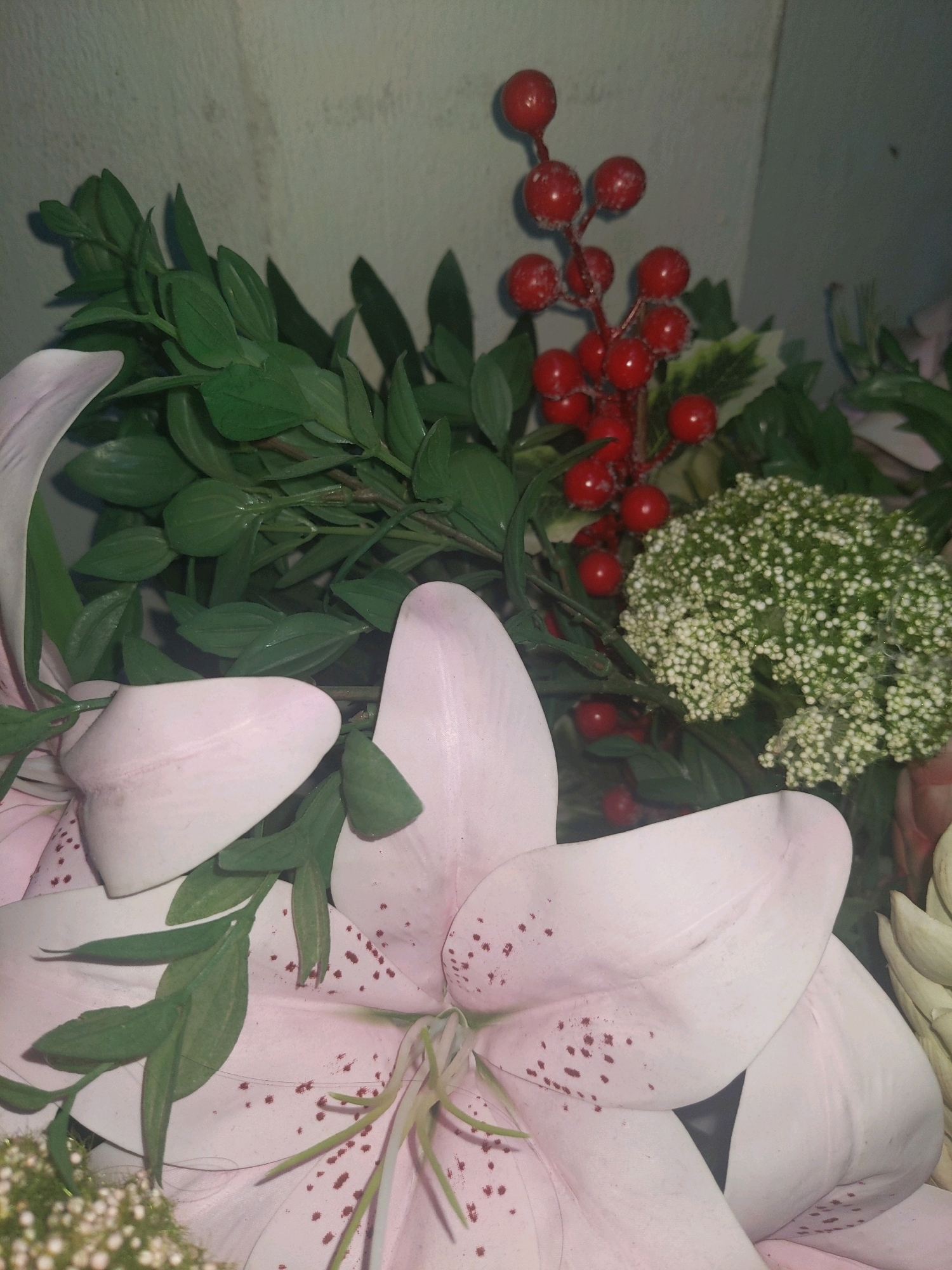 The image shows a close-up of a floral arrangement, possibly for a festive occasion. The arrangement features large, pale pink lilies with speckled petals, interspersed with green foliage. Notable accents include a cluster of small white flowers and a sprig of bright red berries, possibly artificial, with a frosted appearance. The background is a textured, light-colored wall, suggestive of an indoor setting. There are no people or specific geographical landmarks visible to indicate a location in Kolombong, Malaysia, or any other city. The lighting appears to be artificial and somewhat dim, casting subtle shadows on the floral elements.