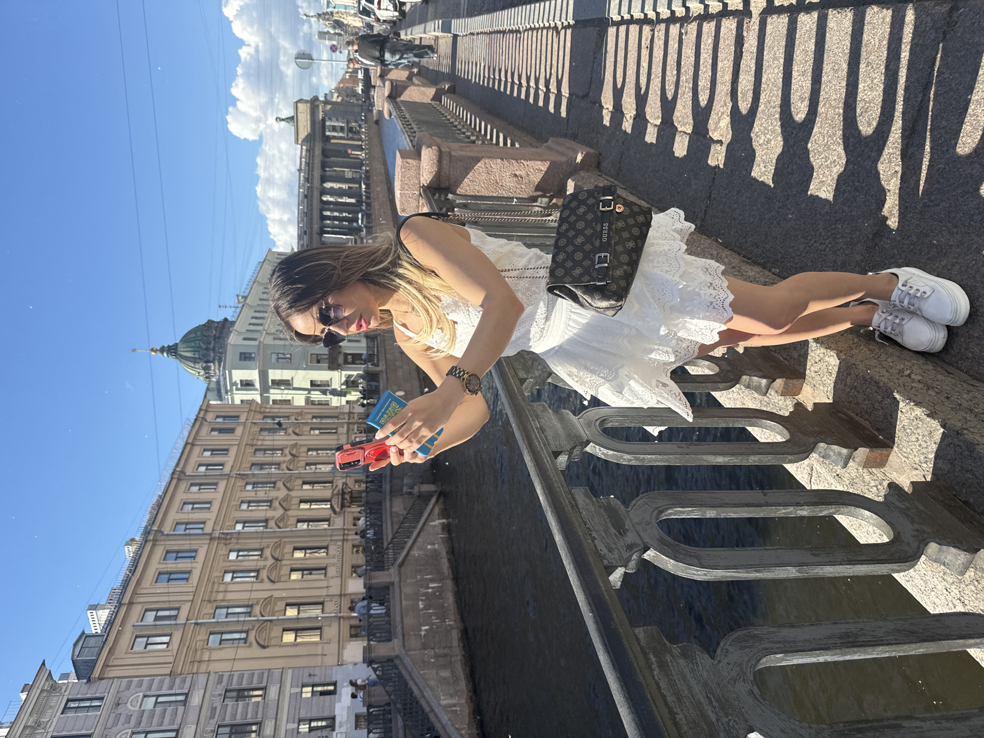 A woman is standing on a bridge overlooking a canal in Saint Petersburg, Russia, holding two phones. She is wearing a white lace dress, sunglasses, and white sneakers. Her purse, a brown Guess handbag, is slung over her shoulder. The canal is lined with stone buildings, and a dome is visible in the distance. The sun is shining, casting shadows from the bridge railing onto the walkway.