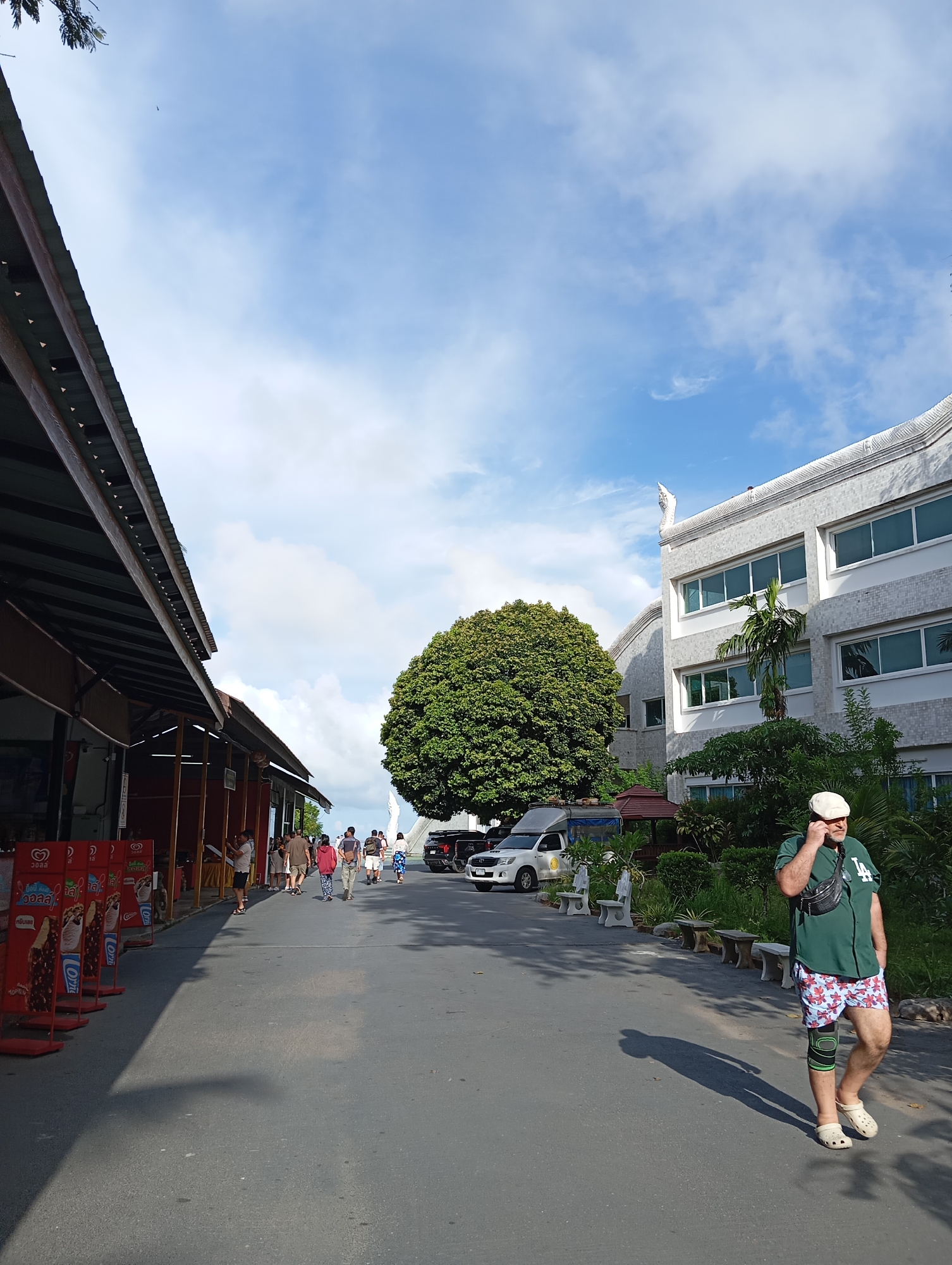 In Karon, Thailand, a man wearing a green baseball jersey, floral-patterned shorts, a knee brace, a white cap, and a black fanny pack walks away from the camera on a paved road, holding a mobile phone to his ear. To his left, a large, round green tree stands prominently. Behind the tree and to the right, a modern, light-colored building with multiple windows is visible. In the foreground to the left, a covered walkway with several ice cream display stands lines the road. Several people are walking in the distance on the road, and a white van is parked further down. The sky is partly cloudy with a bright blue expanse.