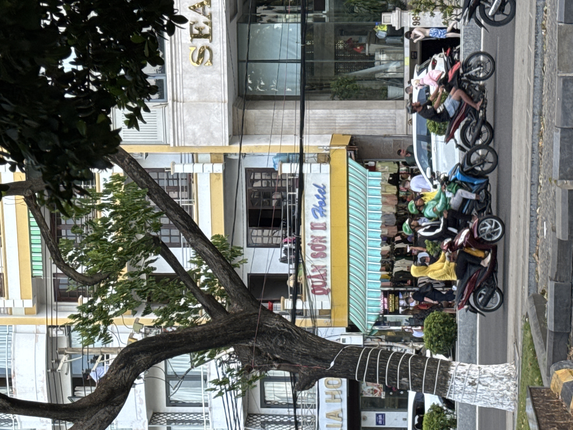 In Nha Trang Ward, Vietnam, a street scene unfolds with a focus on a hotel and surrounding businesses. A tree with a thick, textured trunk and spreading branches dominates the left side of the frame, its white-painted base contrasting with the rough bark. Behind the tree, a multi-story building displays a prominent yellow awning with the words "QUAN SON II Hotel" in stylized pink lettering. Green and white striped awnings extend from the hotel, partially obscuring displays of clothing.
In the foreground, a two-lane road is occupied by several motorbikes. On the left side, a person in a yellow hoodie rides a dark red motorbike. To their right, another person in a green helmet and jacket rides a blue motorbike. Further down the road, a person in a black shirt and blue shorts rides a black motorbike, with a passenger wearing a pink outfit behind them. The motorbikes are in motion, suggesting traffic flow.
Buildings line the street, featuring a mix of architectural styles. One building displays the gold lettering "SEA" at the top. Another has large glass windows reflecting the surroundings. Power lines crisscross the scene above the street and buildings. Pedestrians are visible in the background, including a person in a white shirt standing near a window on an upper level of a building. The overall impression is of a bustling urban environment on a day with diffused light.