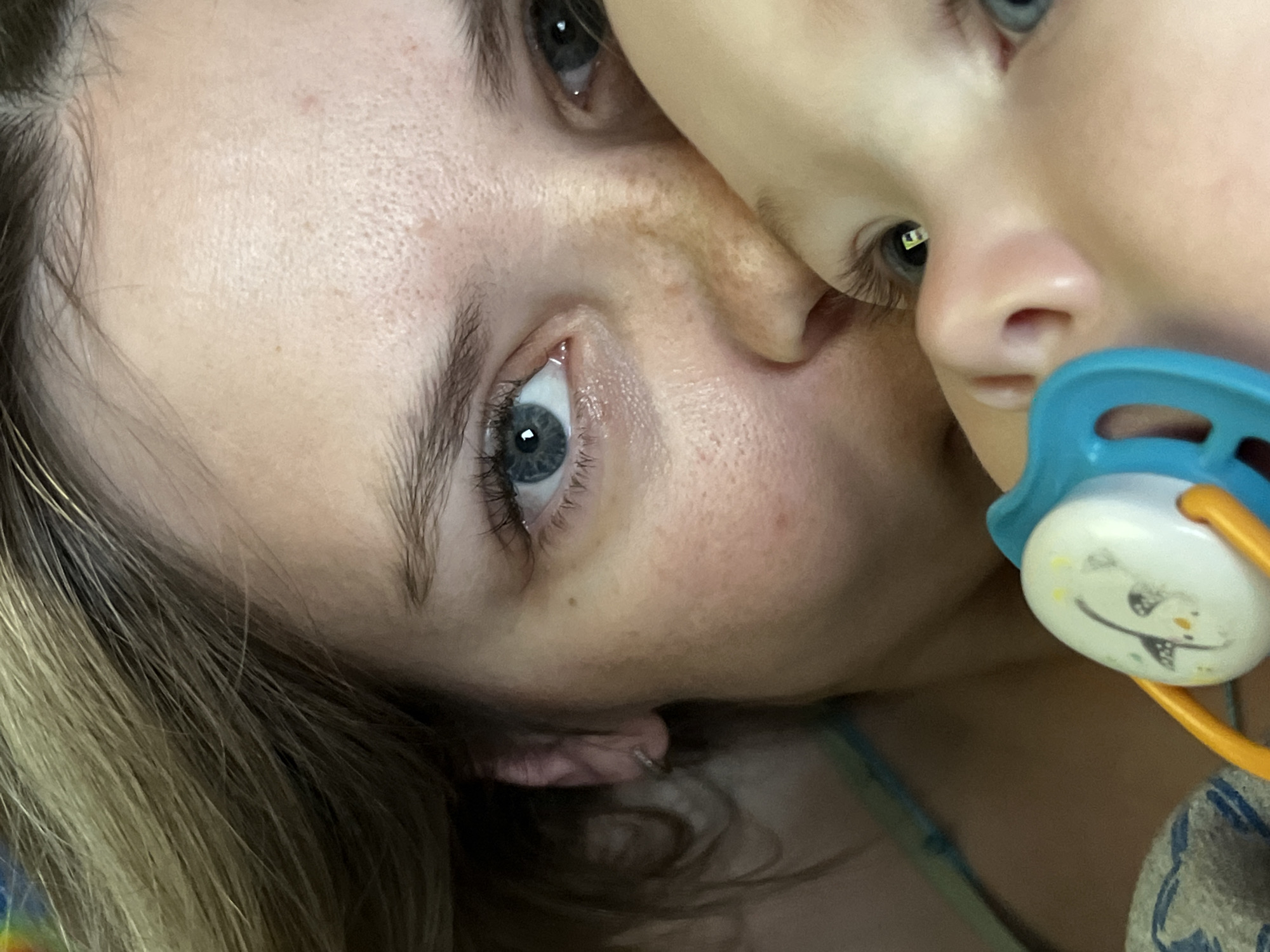 The image shows a close-up of a woman and a child. The woman has blue eyes, light skin, and light-colored hair visible in the corner of the frame. The child is positioned close to the woman, with part of their face visible, including a nose, eye, and eyelashes. The child has a blue and white pacifier with an orange handle near their mouth. The background is out of focus. No landmarks are visible to confirm the location in Chaikovsky, Russia.