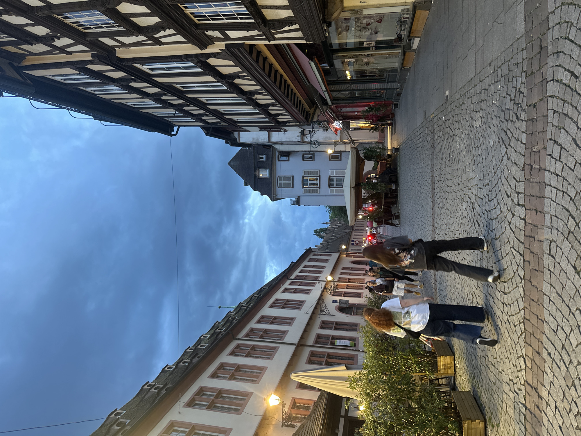 The image depicts a cobblestone pedestrian street in Strasbourg, France, at dusk or early evening. The sky is dark blue and heavily clouded, and streetlights mounted on the buildings are illuminated, casting warm light on the scene.
On the left side of the street, a long, multi-story building with a light-colored facade features reddish-brown window frames and a steeply pitched roof with multiple dormer windows. Along its base, green foliage is arranged in wooden planters, bordering an outdoor seating area with a closed light-colored umbrella.
On the right, a prominent multi-story half-timbered building with dark wooden beams and light-colored infill defines the street edge. Its ground floor houses commercial establishments with red awnings and illuminated display windows. Further down the street, other historic buildings are visible, including a light blue structure with a dark, peaked roof.
Several people populate the street. In the foreground, two women walk away from the camera. The woman on the left has reddish-brown hair, wears a white t-shirt with a graphic, dark wide-leg pants, and carries a dark bag. The woman on the right has dark hair, wears a dark t-shirt with a graphic, dark wide-leg pants, and also carries a dark bag. Other individuals are seen walking further down the street, and some are seated at outdoor tables. A red light, possibly a traffic signal or building light, is visible in the distance. The street itself is paved with grey cobblestones, featuring a stripe of darker, reddish-brown bricks. Overhead wires are visible against the sky.