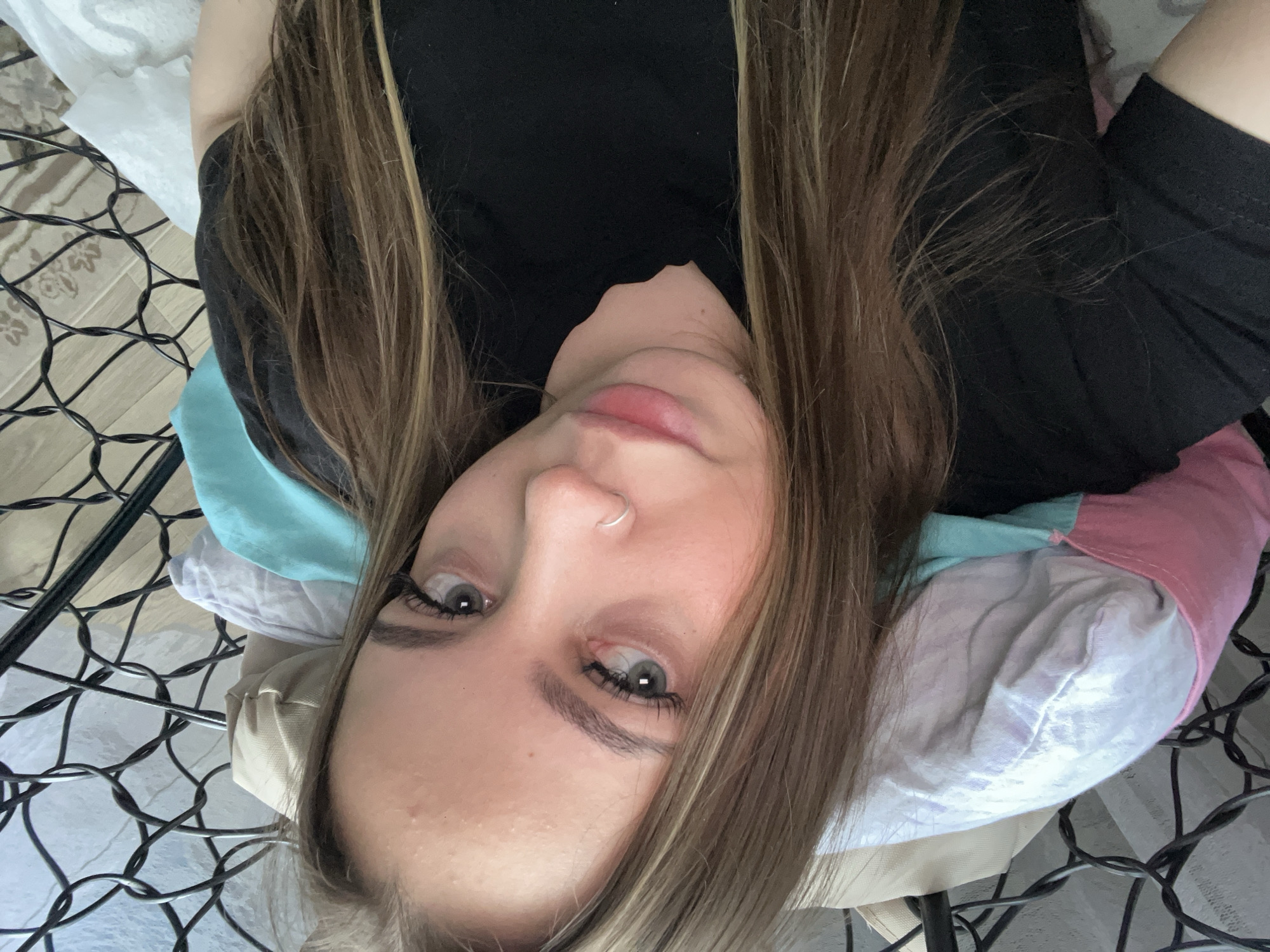 A young woman with long brown hair and a nose ring is lying on a pillow. She is wearing a black shirt. The pillow has multiple colors, including beige, turquoise, white, and pink. The woman is on a metal frame, which appears to be a bed frame. The background is out of focus. There is no evidence in the image to identify the location as Novopokrovskaya, Russia.