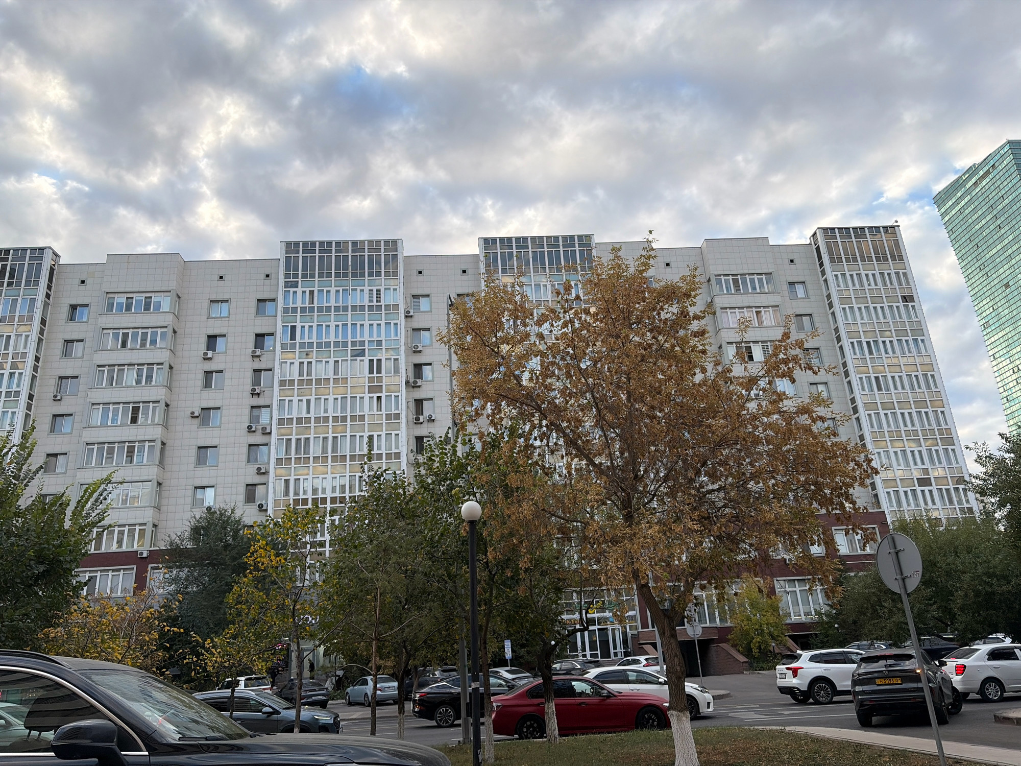 A wide shot captures a street scene in Astana, Kazakhstan, dominated by a large, modern apartment building. The building, with its light-colored facade and numerous windows, stretches across the frame, showcasing a mix of architectural styles, including sections with glass fronts. A taller, modern skyscraper with a distinctive green, glass facade is visible in the background on the right. Several cars are parked along the street in front of the apartment building, including a black SUV and a red sedan in the foreground. Trees with autumn foliage are scattered throughout the scene, adding a touch of color to the urban environment. The sky is overcast with heavy clouds, suggesting a cooler, possibly autumnal, day. The scene appears to be a typical residential area within the city.