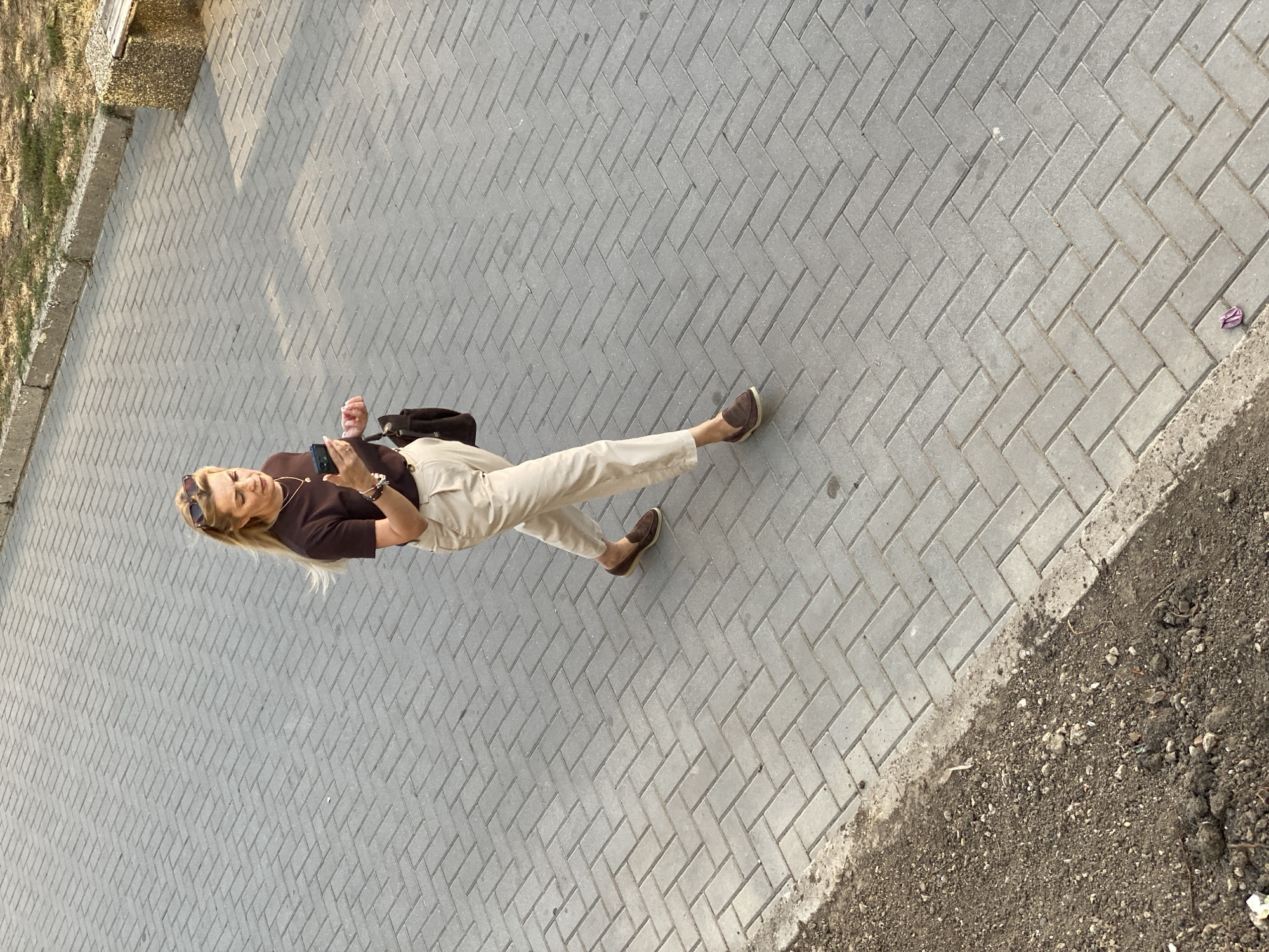 An overhead shot captures a blonde woman walking on a herringbone brick sidewalk in Chișinău, Moldova. She is wearing a dark brown t-shirt, light beige pants, and brown loafers. Sunglasses are perched on her head. She is holding a smartphone in her right hand and a dark purse hangs from her left shoulder. To her right, a patch of dry grass and a concrete bench with a stone base are visible. The sidewalk is bordered by a strip of dirt and gravel on the left. The lighting suggests it is daytime with shadows indicating the sun is positioned to the right.