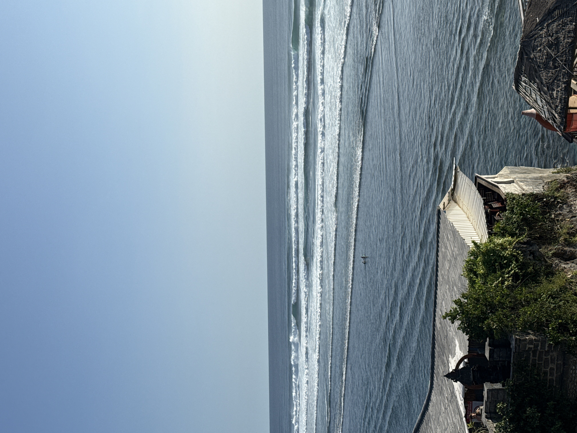 The photograph captures a coastal scene, presumably in Kuta Selatan, Indonesia, given the context. The image is dominated by the ocean, with parallel lines of breaking waves creating a rhythmic pattern on the water's surface. A lone figure is visible in the water.
The right side of the image depicts a rocky coastline with lush vegetation. A structure, possibly a building or temple complex, is perched on the edge of the cliff. The sky is clear and blue, occupying a significant portion of the left side of the photograph.