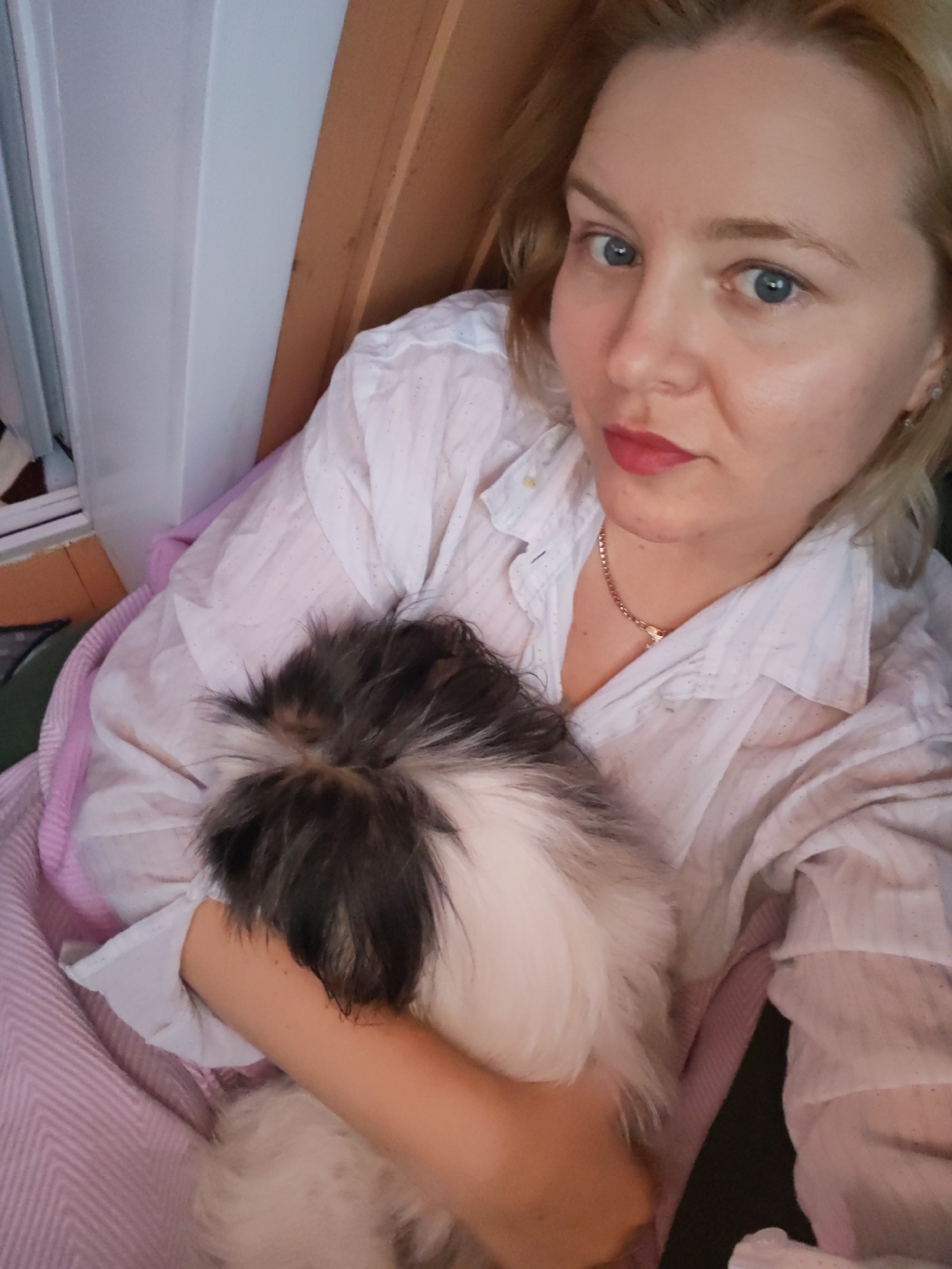 In a medium shot, a blonde woman with blue eyes and pink lipstick is holding a small dog. She is wearing a white collared shirt and a gold chain necklace. The dog is mostly obscured by her arms and the blanket it is wrapped in, but its fur is dark and white. They are indoors, on a couch or bed, with a pink and white striped blanket visible. The background shows a white wall and a wooden panel. This image appears to be a personal photo, taken in a home setting in Izhevsk, Russia.