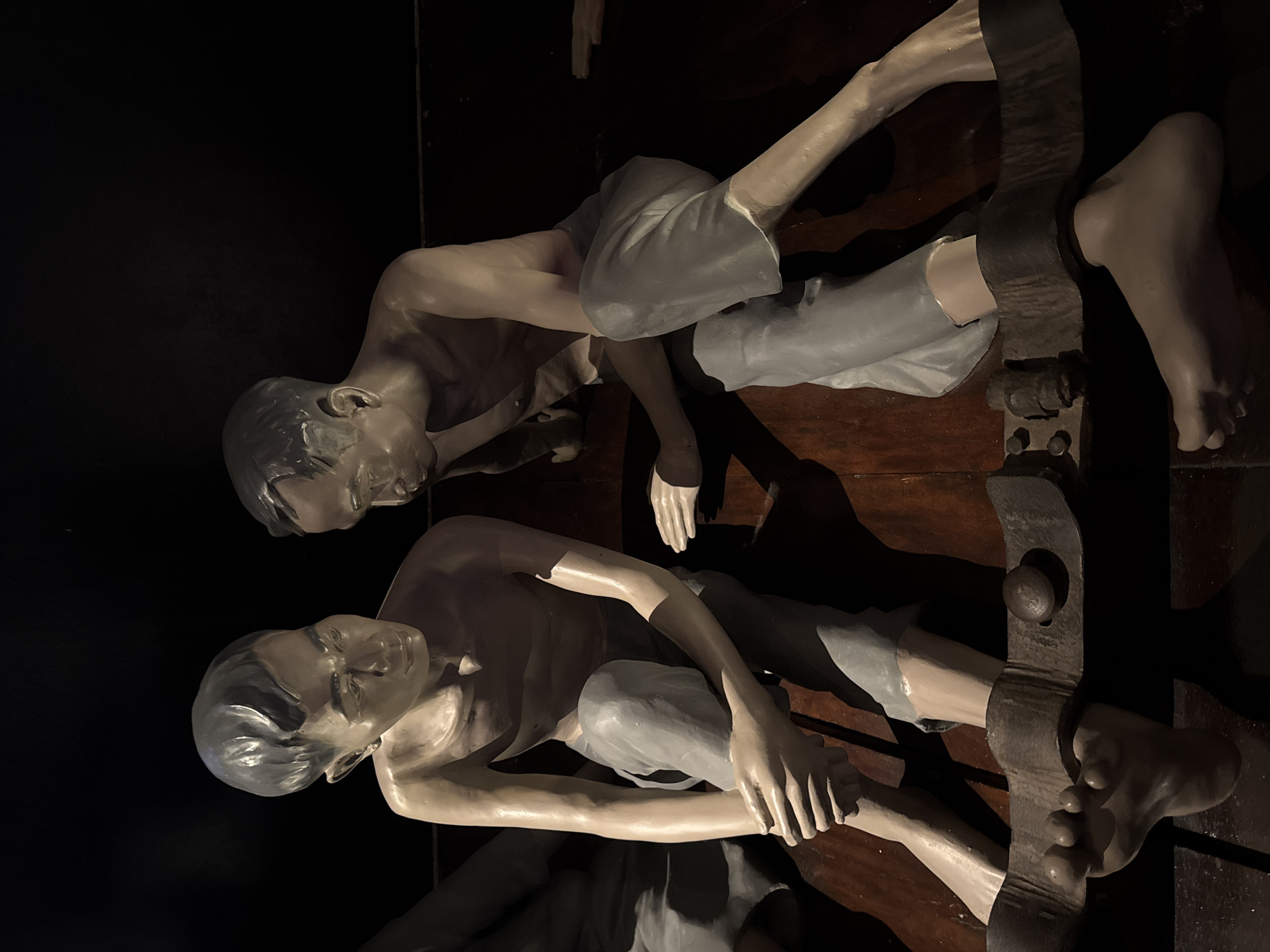 A harrowing exhibit in Hà Nội, Vietnam displays two sculpture figures in a dark enclosure. The figures, dressed only in grey shorts, are fastened to a wooden board by their necks and feet. Their expressions indicate suffering, with eyes downcast and clenched features. The lighting in the enclosure is dim and dramatic, emphasizing the intensity of the scene. The wooden board, likely an instrument of torture, adds to the oppressive atmosphere.