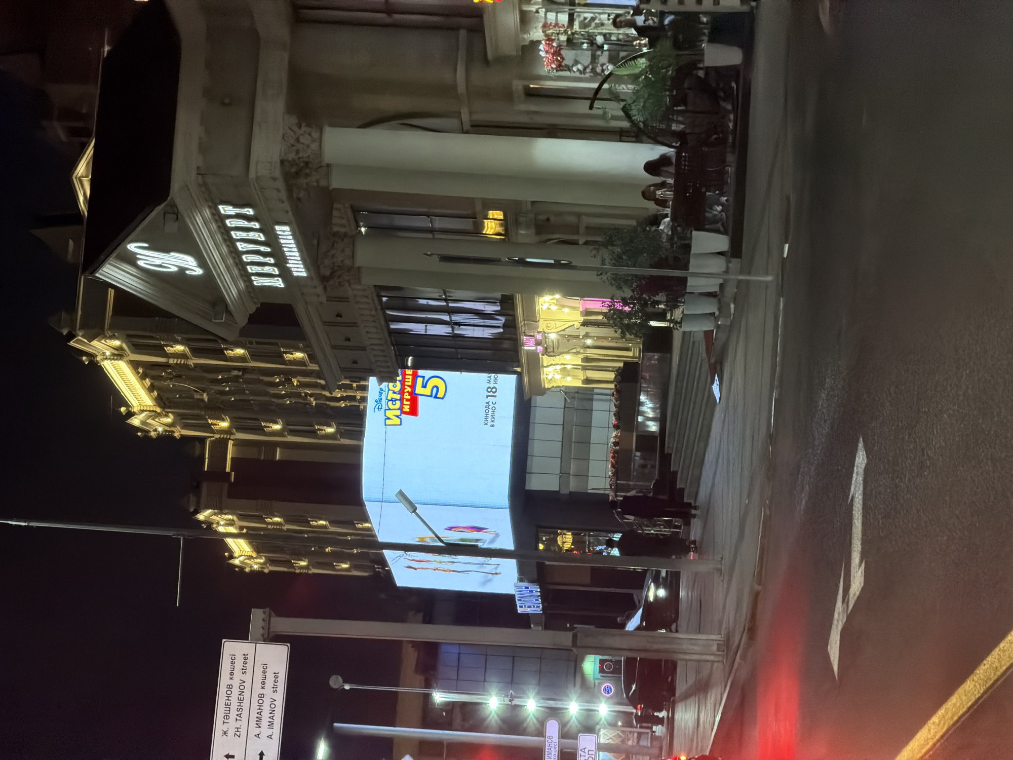 The image captures a street scene at night in Astana, Kazakhstan, specifically on Taushenov street. A large LED screen displays an advertisement for the movie "Toy Story 5" in Russian, indicating a release date of May 18th. Adjacent to the screen is a building with classical architectural elements, featuring columns and ornate detailing, identified by a prominent "M" logo and the word "МЕРУЕРТ" (Meryert) in illuminated lettering. The street is wet, suggesting recent rain, and a car's red taillights are visible on the left side of the frame. People are seen walking and dining at an outdoor seating area of a establishment. Street signs with street names in both Kazakh and Russian languages are also present. The overall ambiance is that of a vibrant urban setting at night, illuminated by artificial lights from buildings and signage.