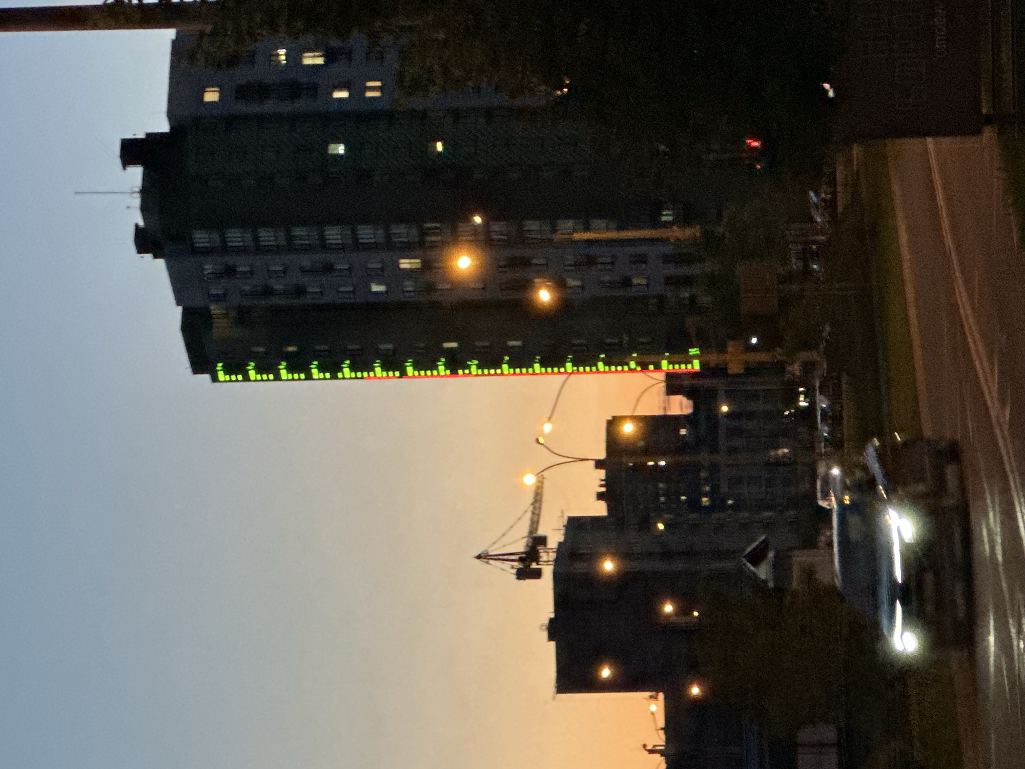 The image captures an urban twilight scene in Minsk, Belarus, looking across a city street towards illuminated buildings. The sky transitions from a light blue-grey at the top to a warm orange-yellow closer to the horizon, indicative of either dusk or dawn.
In the mid-ground on the left, a prominent tall building stands with numerous rectangular windows, some of which are lit with yellow-orange light. A striking vertical line of small, illuminated green and red lights runs down the side of this building. To the right of this, a street stretches with visible lane markings. A car with bright headlights and taillights is captured in motion on the street, indicating ongoing traffic. Streetlights are active, casting pools of warm light onto the road and surrounding areas.
Further into the background, other buildings are silhouetted against the sky, also showing scattered lit windows. A construction crane is visible next to one of these buildings, suggesting urban development. Patches of dark foliage or trees are scattered throughout the scene. On the far right, a dark, vertical structure, possibly a sign or part of a building, shows faint, indistinct white lettering. The overall impression is one of a bustling city transitioning from day to night.