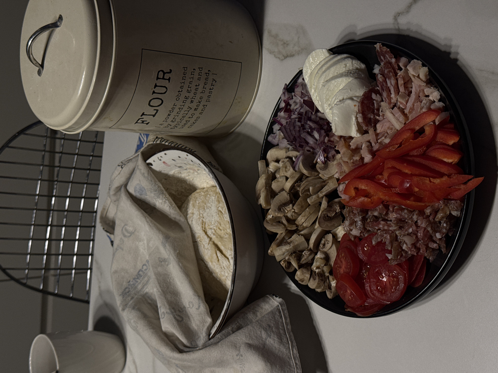 This media file captures a detailed still life of food preparation, likely for baking, set on a kitchen countertop in Ptichnoye, Russia. In the upper left, a cream-colored cylindrical tin with a handle on its lid is prominent. The tin features text including "ΚΟΝΝΠΟΡ" and "FLOUR" in large letters, with a description below: "a powder obtained by grinding grain, typically wheat and used to make bread, cakes and pastry".
To the right, a black circular plate is meticulously arranged with various pizza toppings. These include slices of fresh mozzarella, different types of cured meats such as sliced salami and shredded chicken or ham, vibrant red bell pepper strips, sliced cherry tomatoes, sliced mushrooms, and finely diced red onion.
In the lower-left, a white bowl holds prepared dough, partially covered by a light-colored, possibly flour-dusted, fabric cloth or towel. A metal cooling rack stands vertically behind the bowl, and a white ceramic mug is partially visible below the rack. The lighting is somewhat dim, casting subtle shadows, suggesting an indoor setting, possibly during the evening. The overall scene conveys a sense of organized culinary activity.