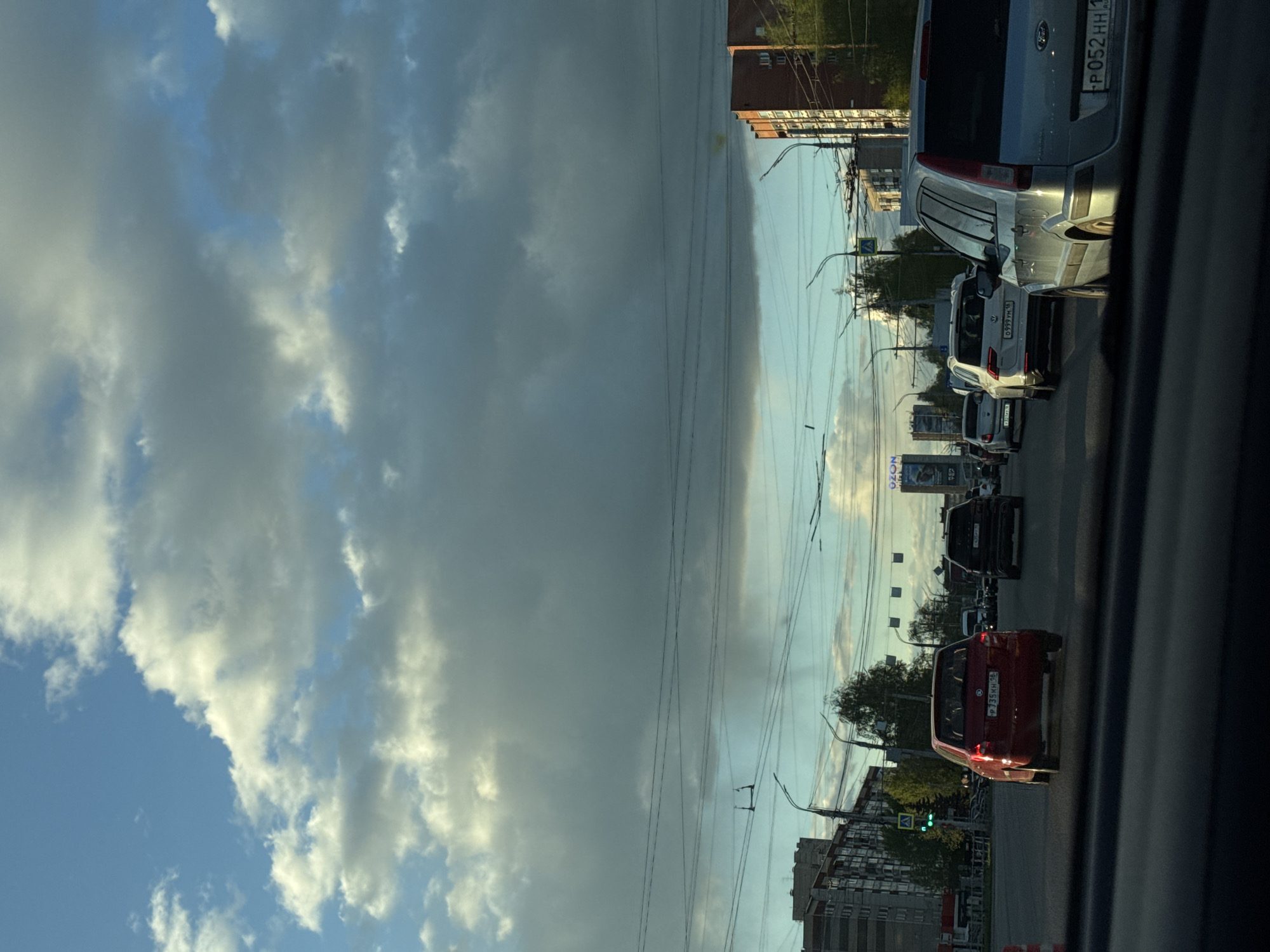The image shows a street scene in Izhevsk, Russia. The sky is partially cloudy. A line of cars is visible, and the license plates on the vehicles can be seen. Utility poles with power lines line the street. Buildings of varying sizes are in the background. A billboard and traffic lights are visible along the side of the road. The photograph was likely taken from inside a car.