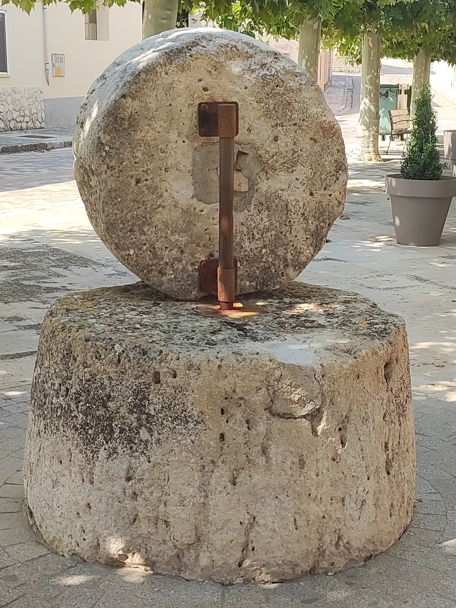 The image shows an outdoor scene in Ampudia, Spain, featuring an old millstone displayed as a monument. The millstone consists of two large, rough-hewn stones, one stacked on top of the other. A vertical metal rod with a horizontal piece attached runs through the center of the upper millstone, securing it to the base. The stones are weathered and pitted, with visible texture and imperfections.
The millstone is placed on a paved area, possibly a town square or walkway, with patterned grey stones. In the background, there are several large trees with lush green foliage, casting dappled shadows on the ground. Beyond the trees, a building with white walls and windows is visible, along with a street scene that includes a bench, a trash receptacle, and a potted plant with a small evergreen shrub. The lighting suggests a sunny day, with strong sunlight and shadows. The overall environment appears to be a public space within a town, likely a historical or cultural point of interest.