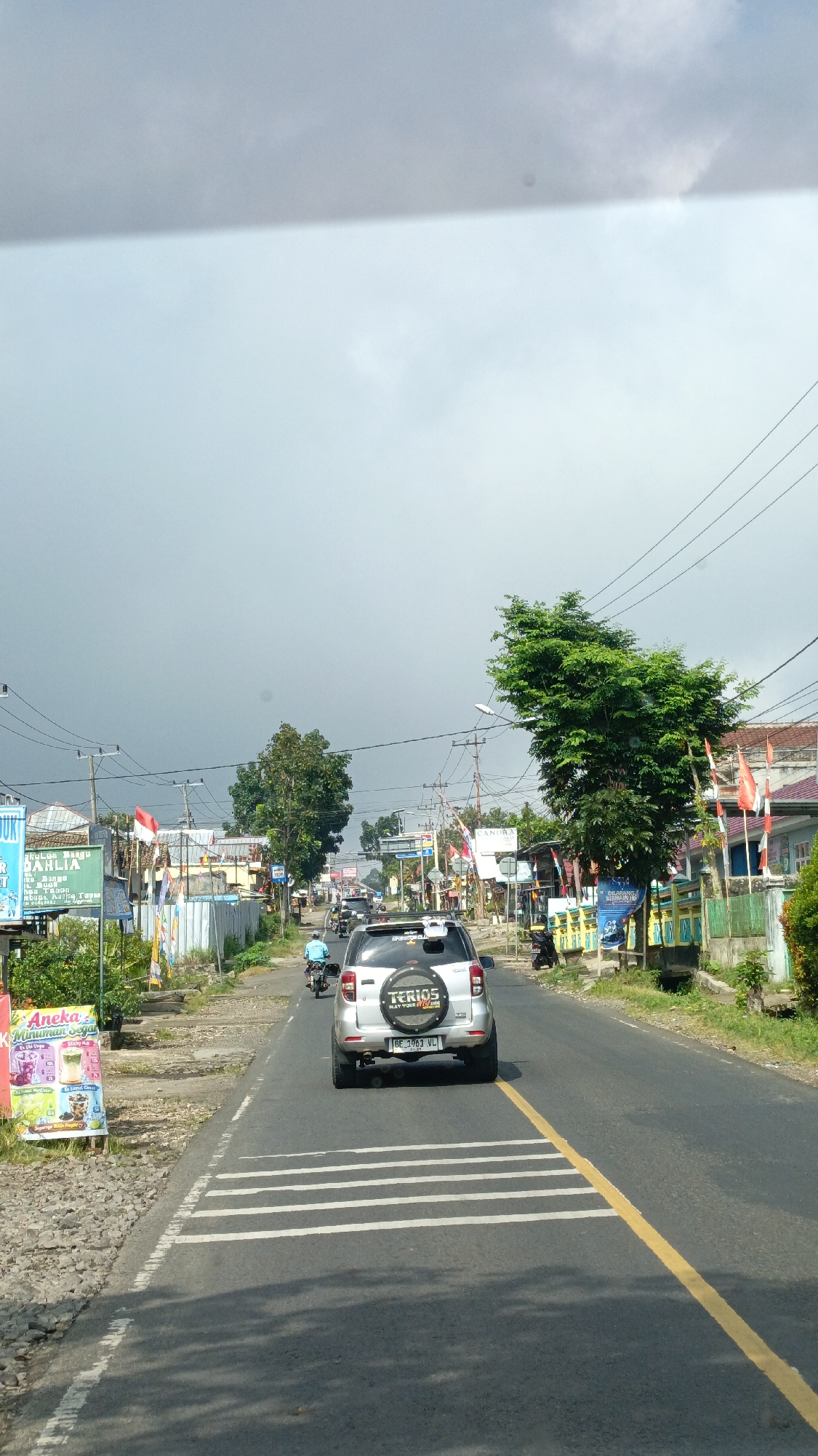 The image shows a street scene in Sukabumi, Indonesia. The viewpoint is from a vehicle looking forward. The road is paved with asphalt and has a yellow center line and white crosswalk markings. Several vehicles, including a silver SUV with "TERIOS" written on the back and a motorcycle with a rider in a blue shirt, are traveling on the road. On the left side of the road, there are buildings, signs, and vegetation. One sign reads "Aneka Minuman Segar". On the right side of the road, there are more buildings, flags (some appearing to be Indonesian flags), and trees. The sky is overcast with some lighter patches of sky visible. The overall impression is of a moderately busy street in a town or city.
