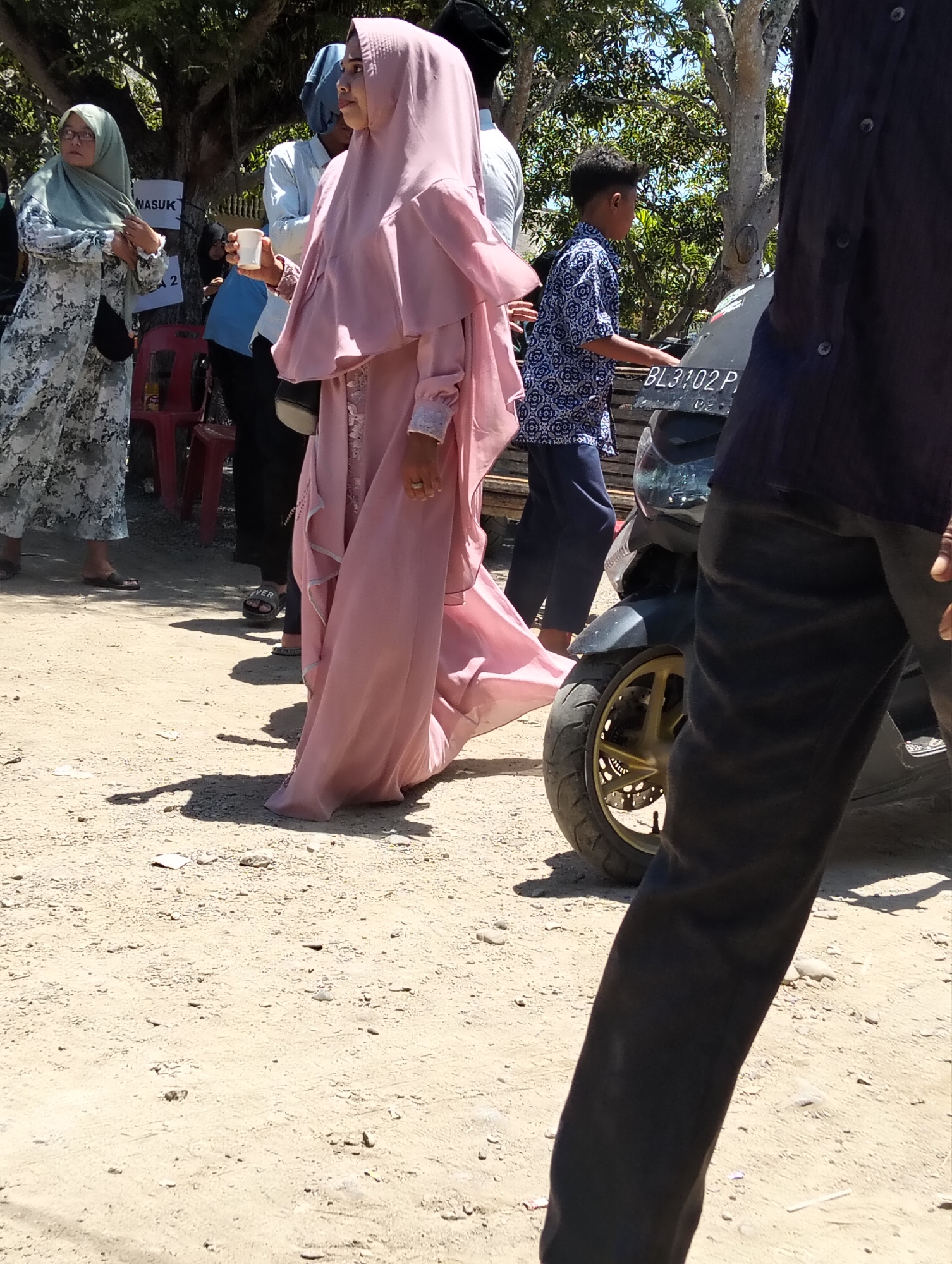 In Teungoh, Indonesia, on a sunny day, a group of people are gathered outdoors. The foreground is dominated by the legs of a person wearing dark trousers, partially obscuring a black motorcycle with a gold-spoked wheel and a license plate reading "BL 3102 P". A young boy in a blue patterned shirt and dark trousers is visible near the motorcycle, his left hand extended. To the left, a woman in a long pink hijab and dress is walking away from the camera, holding a white cup. Another woman to her left wears a light green hijab and a patterned dress. In the background, a sign with "MASUK" and "2" is visible, and red chairs are present. The ground is covered in light-colored gravel and dirt, with scattered debris and shadows indicating the time of day. Trees with dense foliage provide shade in the background.