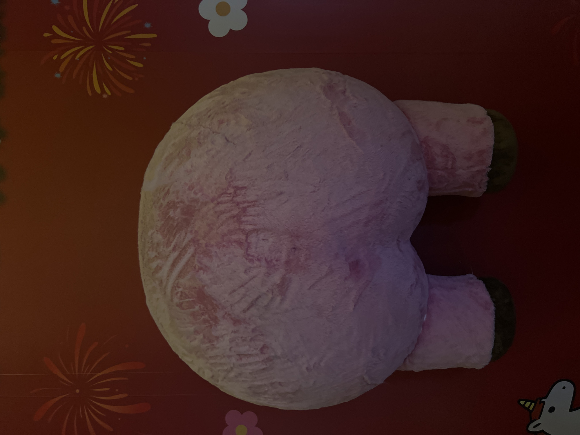 A pink plush toy with brown hooves is centered in the image, viewed from behind. The background is a reddish-brown surface with cartoon-like decorations including fireworks, flowers, and a unicorn. There is no evidence to indicate that the image was taken in Futian District, China.