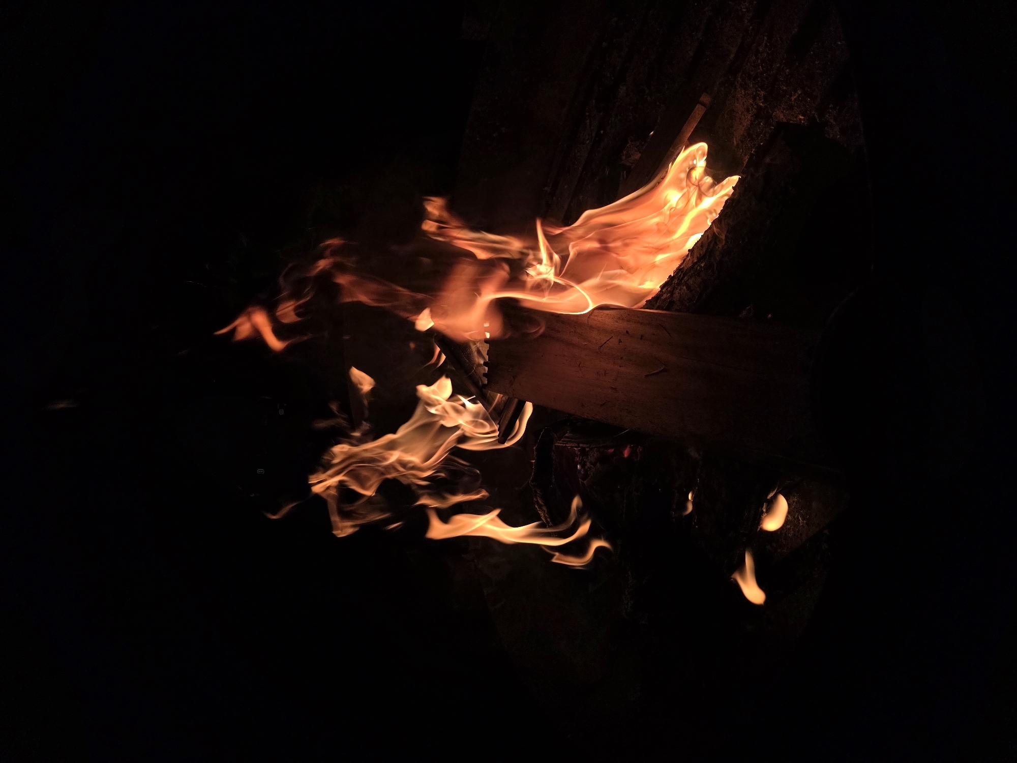 A campfire burns at night in a dark, outdoor setting. The flames are a vibrant orange and yellow, illuminating a piece of firewood and surrounding embers. The background is almost entirely black, creating a stark contrast with the bright fire. There are no people visible in the image. The scene appears to be in a natural or semi-natural environment, possibly a backyard or a designated fire pit area. The location is specified as Строитель-нефтяник Снт, Russia.
