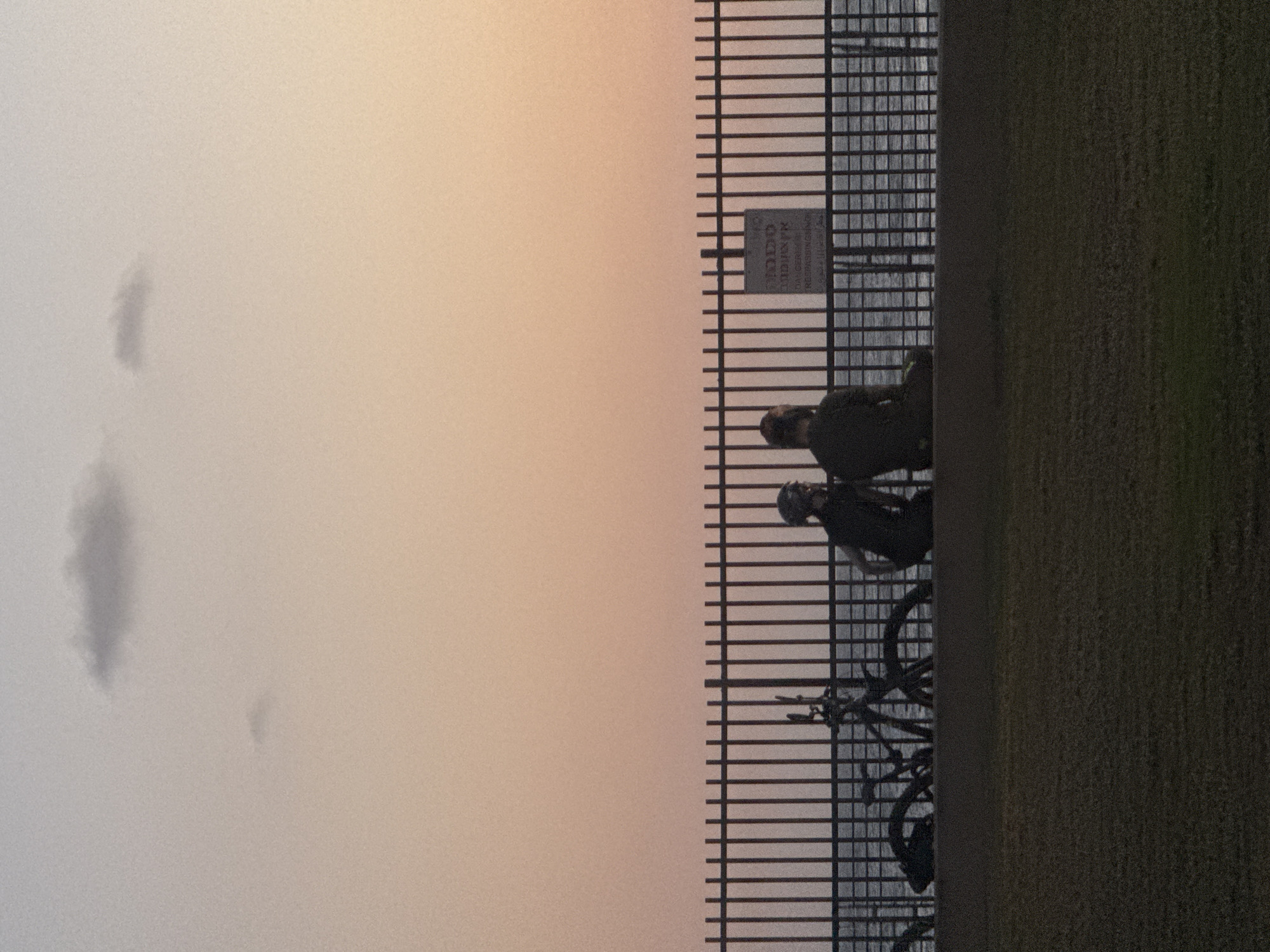 The image depicts a scene along a fence, possibly a waterfront, in Tel Aviv, Israel. Two people are near the fence with bicycles. The fence is made of vertical metal bars. Beyond the fence, there is a body of water, likely the Mediterranean Sea. To the right, there is a dark, textured surface, potentially a wall or grassy area. The sky is a pale, dusky color, possibly indicating twilight or a cloudy day. The lighting is subdued.