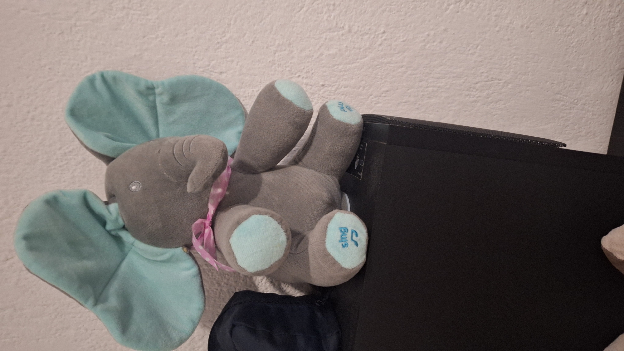 A grey plush elephant toy with light blue ears and footpads lies on its side, leaning against a dark rectangular object. The elephant features a pink ribbon tied around its neck and small, shiny plastic eyes. On one of its light blue footpads, the stylized word "sing" is visible, suggesting it might be an interactive toy. The background consists of a textured, off-white wall. Part of a dark blue bag is visible beneath the elephant. The dark rectangular object it leans against appears to be an electronic device, possibly a speaker or computer component. The scene is an indoor setting in Blagoevgrad, Bulgaria, under ambient artificial or natural light, though the exact time of day is not discernible. No specific activity is taking place beyond the stationary arrangement of the objects.