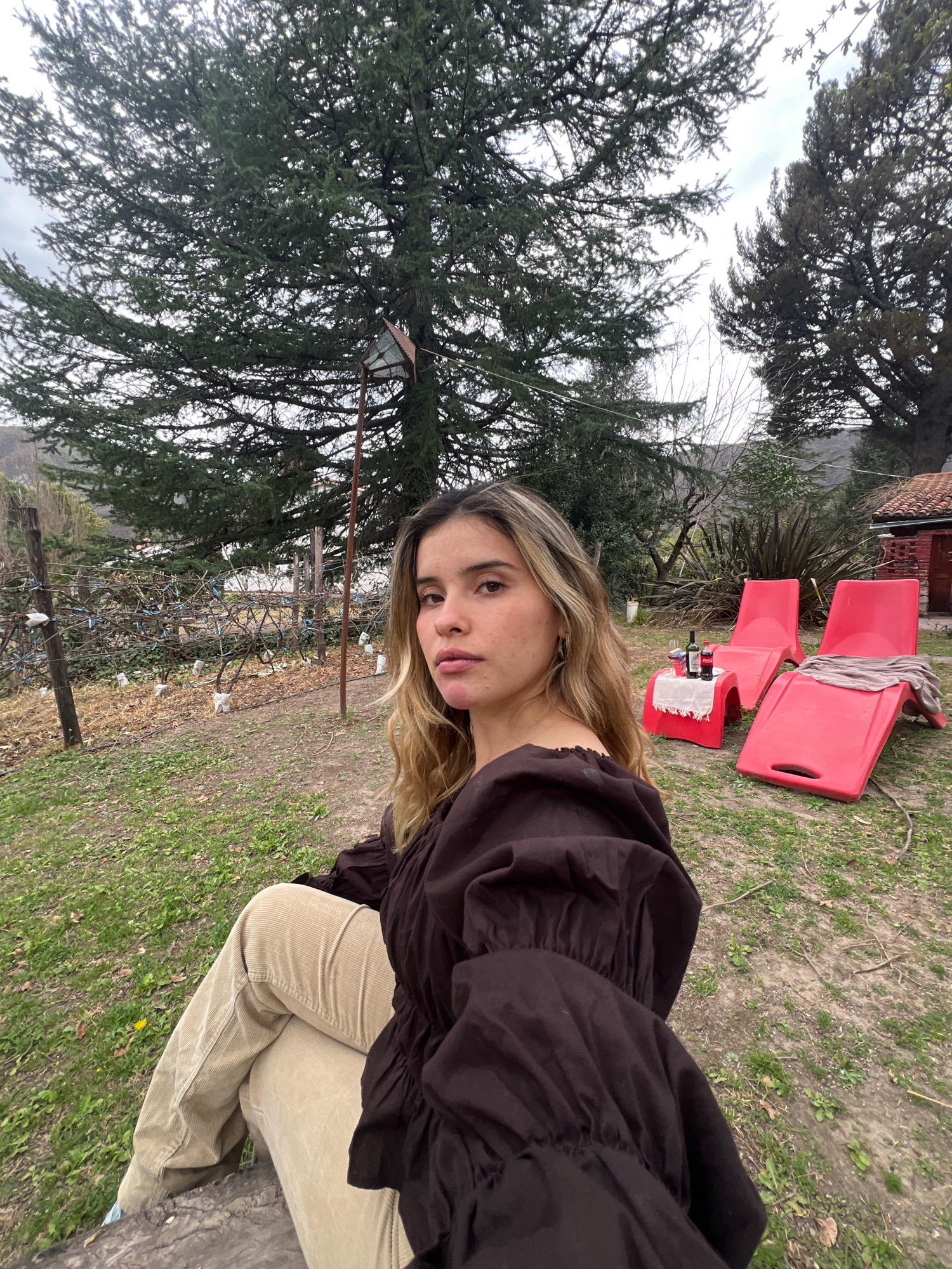 In El Rodeo, Argentina, a young woman with blonde hair sits on a wooden surface in an outdoor setting. She is wearing a dark brown puffed-sleeve top and light brown corduroy pants. She is looking towards the camera. In the background, there are two red lounge chairs and a small table with a bottle of wine and glasses. A tall tree with dark green foliage stands behind the woman. The ground is covered with grass and fallen leaves. A rustic fence made of wooden posts and wire runs along the left side of the image. The sky is overcast.