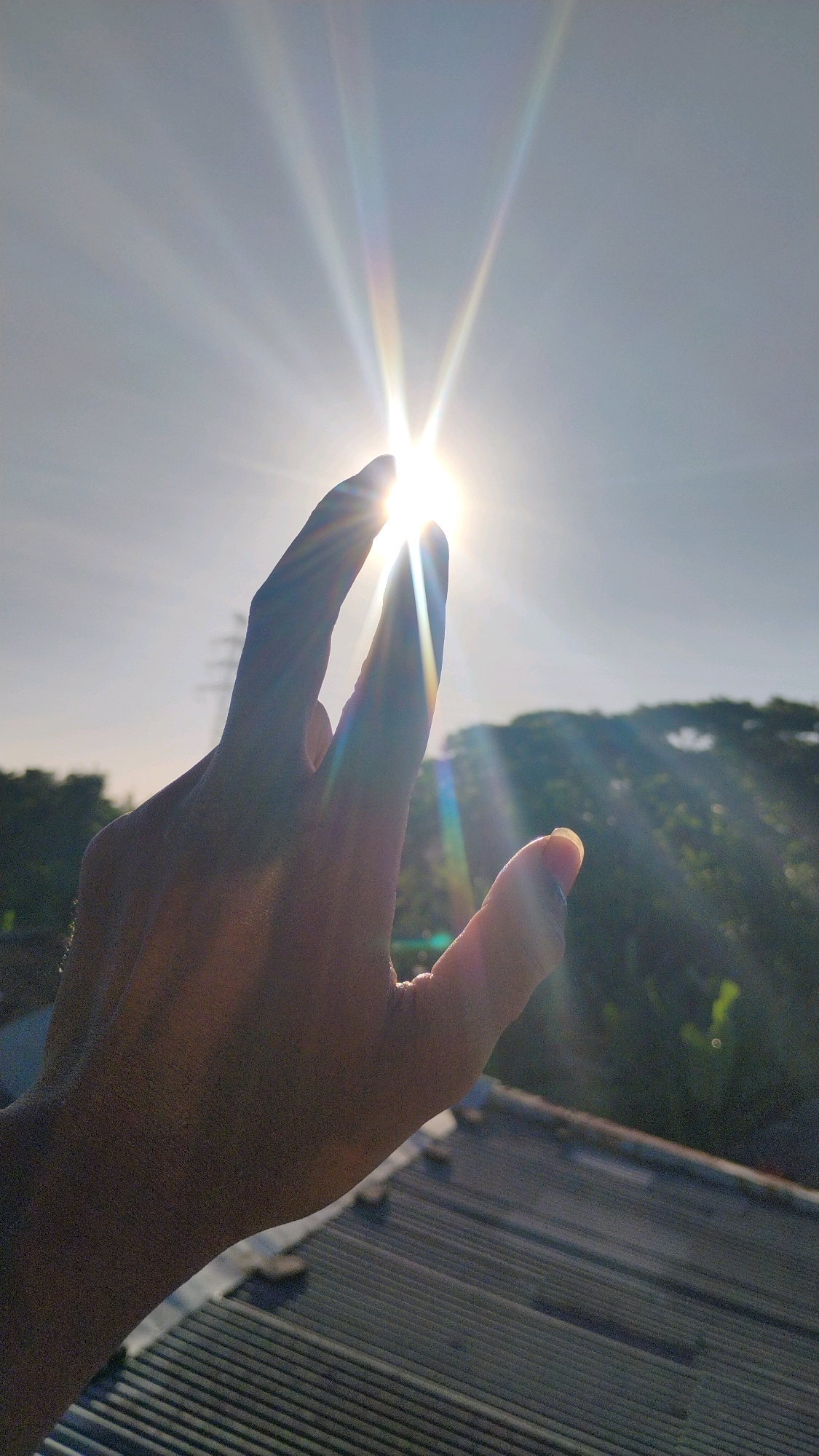 A human hand is held up, its fingers extended to block the bright sun. The sun's rays refract, creating a rainbow effect. In the background, a cityscape of Gumpang, Indonesia, is visible, with rooftops and trees bathed in sunlight. The sky is clear and bright blue.