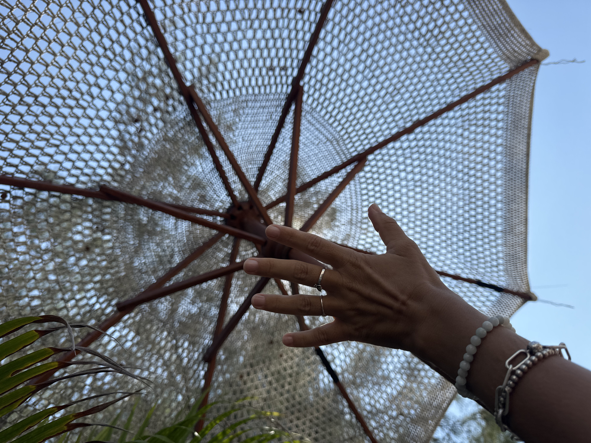 An upward-facing, close-up shot shows a person's hand reaching up towards a woven umbrella canopy. The umbrella, with its wooden spokes, is stretched open against a bright blue sky. The hand, adorned with two rings on its fingers, is held open, palm facing upwards. A beaded bracelet and a chunky silver bracelet are visible on the person's wrist. Green palm fronds are visible in the upper left corner of the frame. The image appears to be taken outdoors, likely in a tropical or beach setting.
